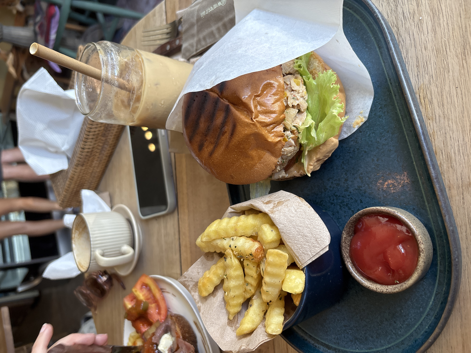 A table at a restaurant in Kuta Selatan, Indonesia, holds a tray with a burger wrapped in paper, a small dish of french fries, and a dish of ketchup. A glass of iced coffee with a straw sits next to the tray, along with a coffee cup and a plate with food. A smartphone is also on the table. Parts of people and chairs are visible in the background.
