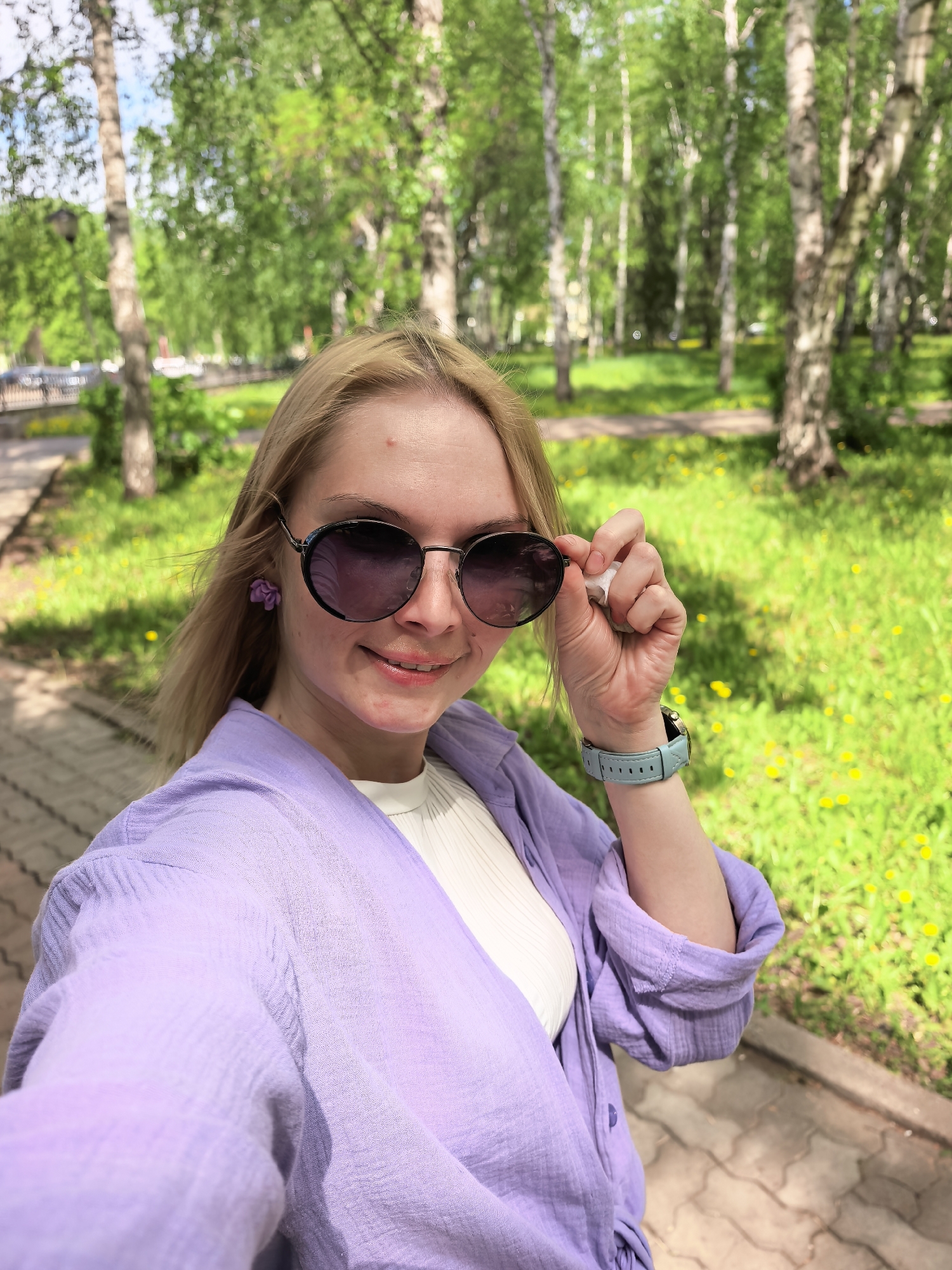 A young woman stands on a paved path in a park setting. She is fair-skinned with blonde hair and wearing sunglasses, a lavender-colored blouse, and a white shirt underneath. She is holding her sunglasses slightly away from her face with her right hand. On her left wrist, she wears a light blue smartwatch. The background shows a grassy area with yellow flowers, birch trees, and other trees. The location is Sterlitamak, Russia.