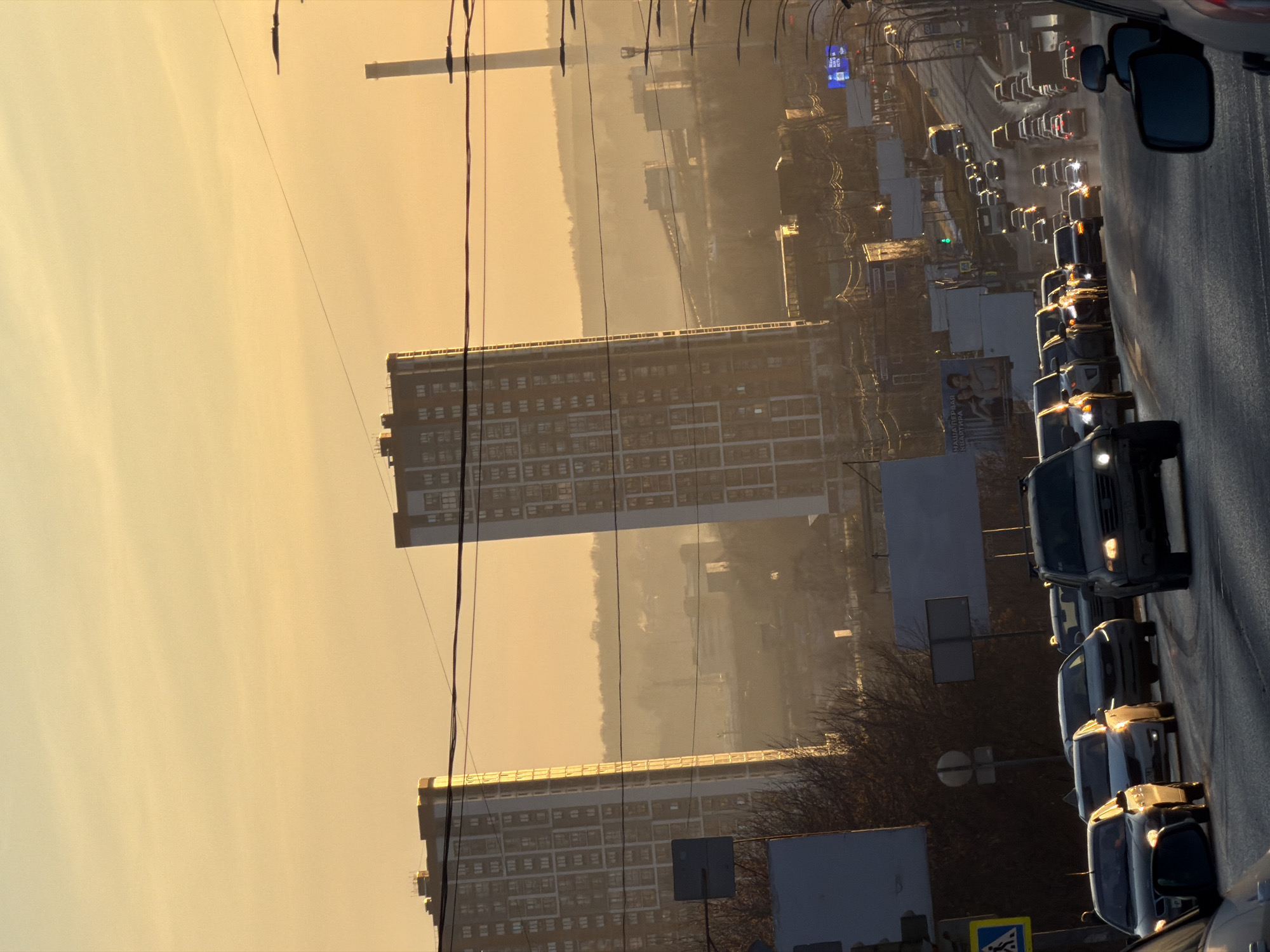 The image depicts a traffic jam on a road in Izhevsk, Russia. Cars are lined up on the right side of the frame, with a focus on a dark-colored minivan leading the line. In the background, there are tall apartment buildings with repetitive window patterns. Power lines are visible in the sky. The atmospheric conditions suggest either sunset or sunrise, casting a yellow/orange hue over the sky and cityscape. A billboard and various traffic signs are also visible along the roadside.