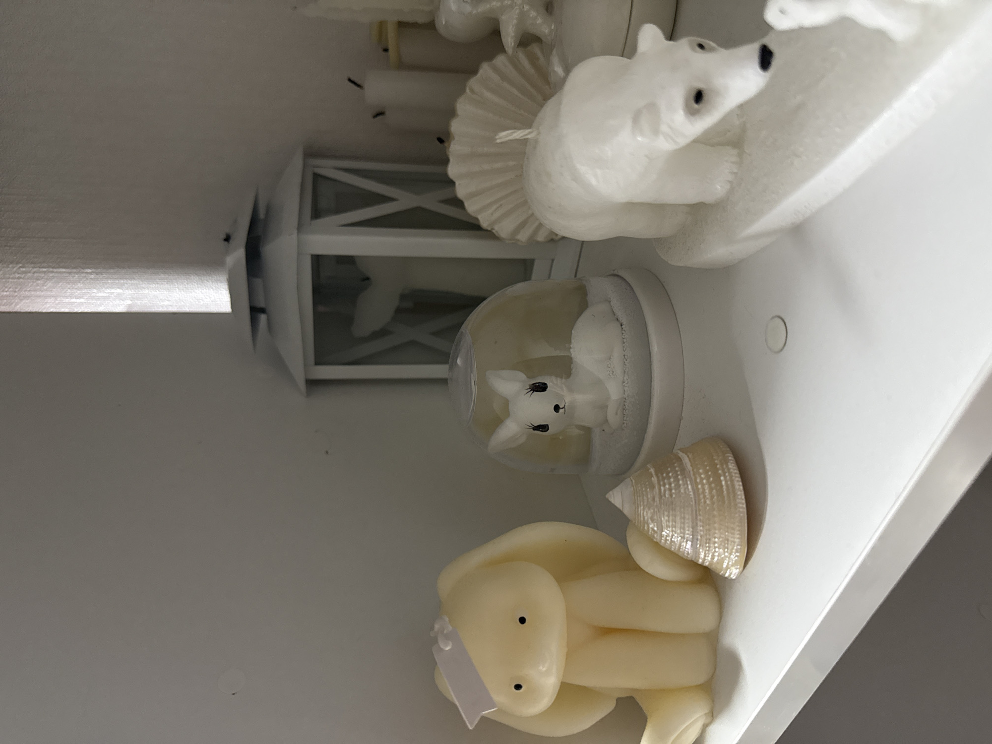 The image depicts an indoor scene in the city of Vniissok, Russia, showcasing a collection of decorative objects, primarily candles, arranged on a white shelf against a plain, light-colored wall. The setting is illuminated by soft, diffuse lighting.
On the left side of the white shelf, a cream-colored candle shaped like a sitting rabbit or bunny is present. This candle features small black eyes and a small, white rectangular tag attached to its head with a thin string. To its immediate right, a small, light-toned conical seashell with iridescent markings rests on the shelf. Adjacent and slightly behind, a clear glass dome encloses a white figurine of a sitting deer, detailed with small black eyes and lashes, which stands on a textured white base resembling snow.
In the mid-ground, a white decorative lantern with a pointed, tiered roof and white cross-patterned window frames is positioned. A soft light source is visible inside or behind the lantern, casting a faint glow. Inside the lantern, a light-colored object, possibly a candle sculpted as a whale tail, is discernible. To the right of the lantern, a cluster of several plain white pillar candles of varying heights is arranged; one appears partially consumed with a darkened wick.
In the right foreground, a white candle sculpted as a standing polar bear is visible, featuring small black eyes and a black nose, resting on a flat white base. A second, smaller white shape, possibly a polar bear cub or another decorative element, is partially visible in the far right foreground, also on a white base. Behind the prominent polar bear, a large, light-colored, fan-shaped object, resembling a shell, stands upright.
The white shelf surface exhibits several small, faint circular indentations. The background wall is uniformly light-colored, possibly off-white, and shows a subtle vertical texture, particularly in the upper right section of the frame.