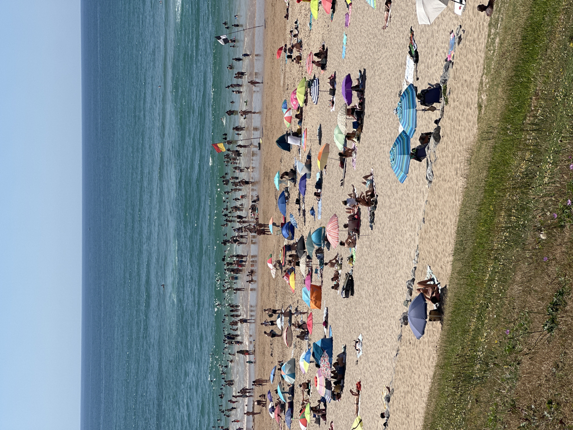 This is a photograph of a crowded beach in Biarritz, France, on a sunny day. The ocean is a vibrant blue, with small waves. Many people are in the water, swimming and playing. On the sandy beach, numerous umbrellas provide shade for sunbathers. People are scattered across the sand, some sitting, some lying down, and some walking. Beach tents are also visible. In the background, a red and yellow flag and a black and white flag are planted in the sand, likely indicating lifeguard stations or designated swimming areas. The overall scene depicts a lively and popular beach resort.