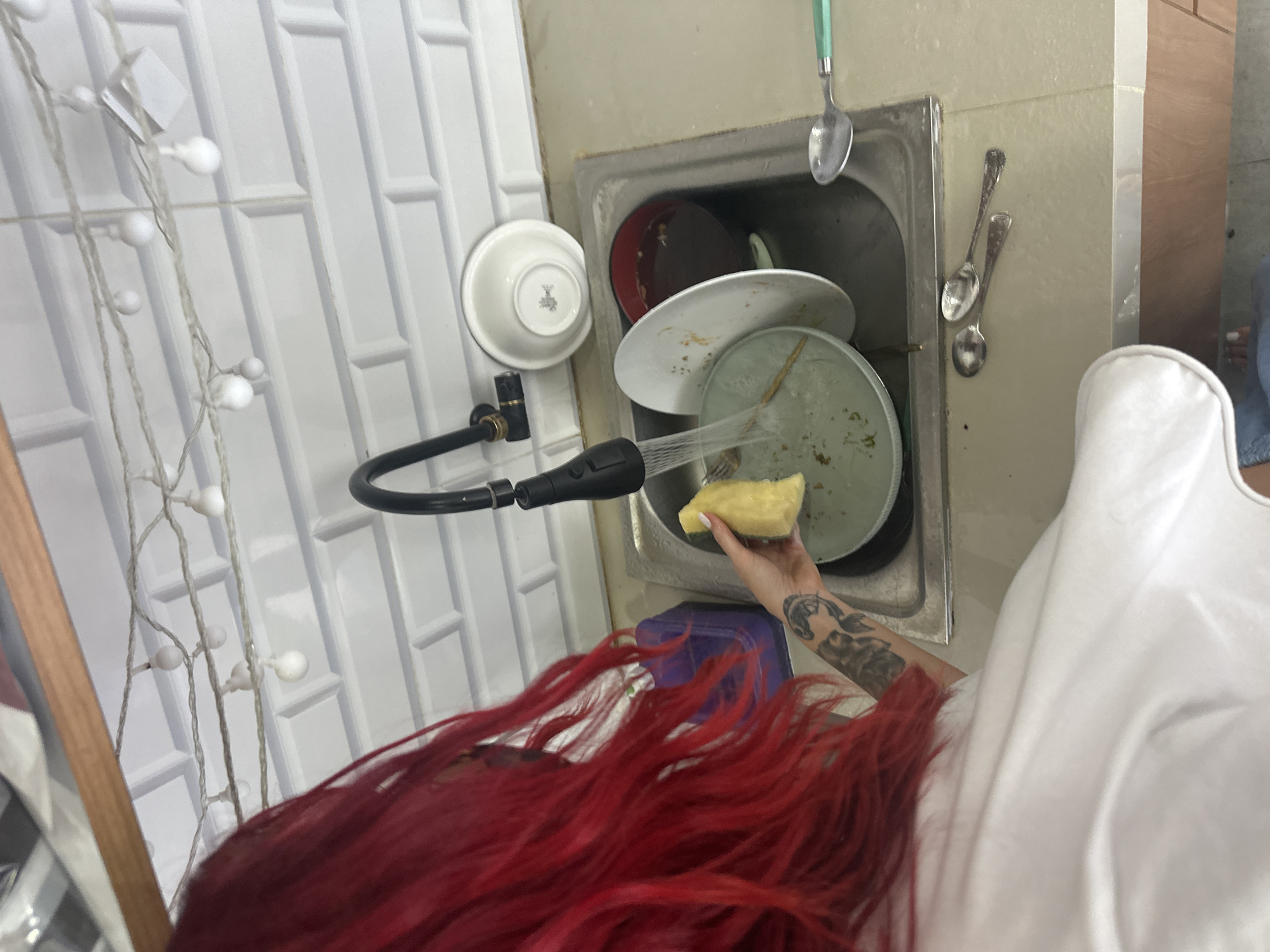 An overhead, vertically oriented photograph captures a person with long, vibrant red hair and a white top engaged in dishwashing. The individual's right arm, adorned with tattoos, extends into a metal kitchen sink, holding a yellow sponge. Water sprays from a black faucet attachment onto a stack of dirty dishes, which includes several plates and bowls showing food remnants. A red bowl is visible among the stacked items.
Above the sink, a spoon with a light green handle hangs, while two other spoons are affixed to the wall next to the sink. The wall to the left of the sink is covered with white, rectangular subway-style tiles, from which a string of white, spherical fairy lights is draped. Part of a wooden frame is visible on the far left of the image. The setting appears to be an indoor kitchen or utility area, with sufficient ambient light to suggest daytime. A purple plastic container is partially visible beneath the sink. No specific external location cues are present in the image itself, though the context indicates North Kuta, Indonesia. No readable text or discernible emotions are visible. The scene depicts a routine domestic activity.