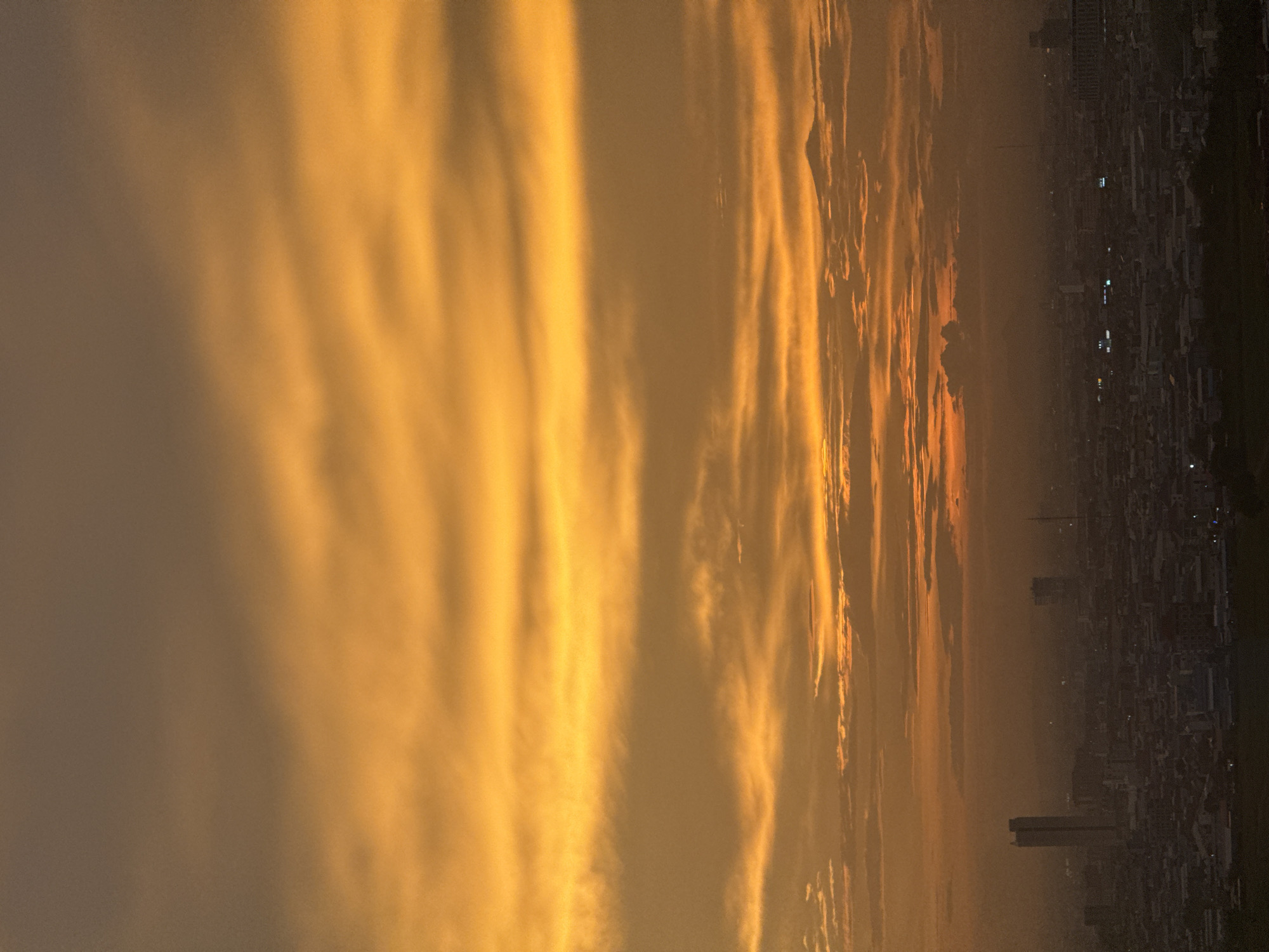 The image captures a cityscape, likely taken from an elevated vantage point, under a dramatic sunset. The sky is dominated by vibrant hues of orange and yellow, with distinct horizontal cloud formations. In the middle ground, a dense urban sprawl is visible, characterized by a multitude of buildings and structures. A tall, slender skyscraper stands out on the left side of the frame. The lower portion of the image shows more buildings and what appears to be a natural landscape or undeveloped area in the foreground. The atmosphere appears hazy, possibly due to pollution or the time of day. The scene depicts a moment of natural beauty in an urban environment.