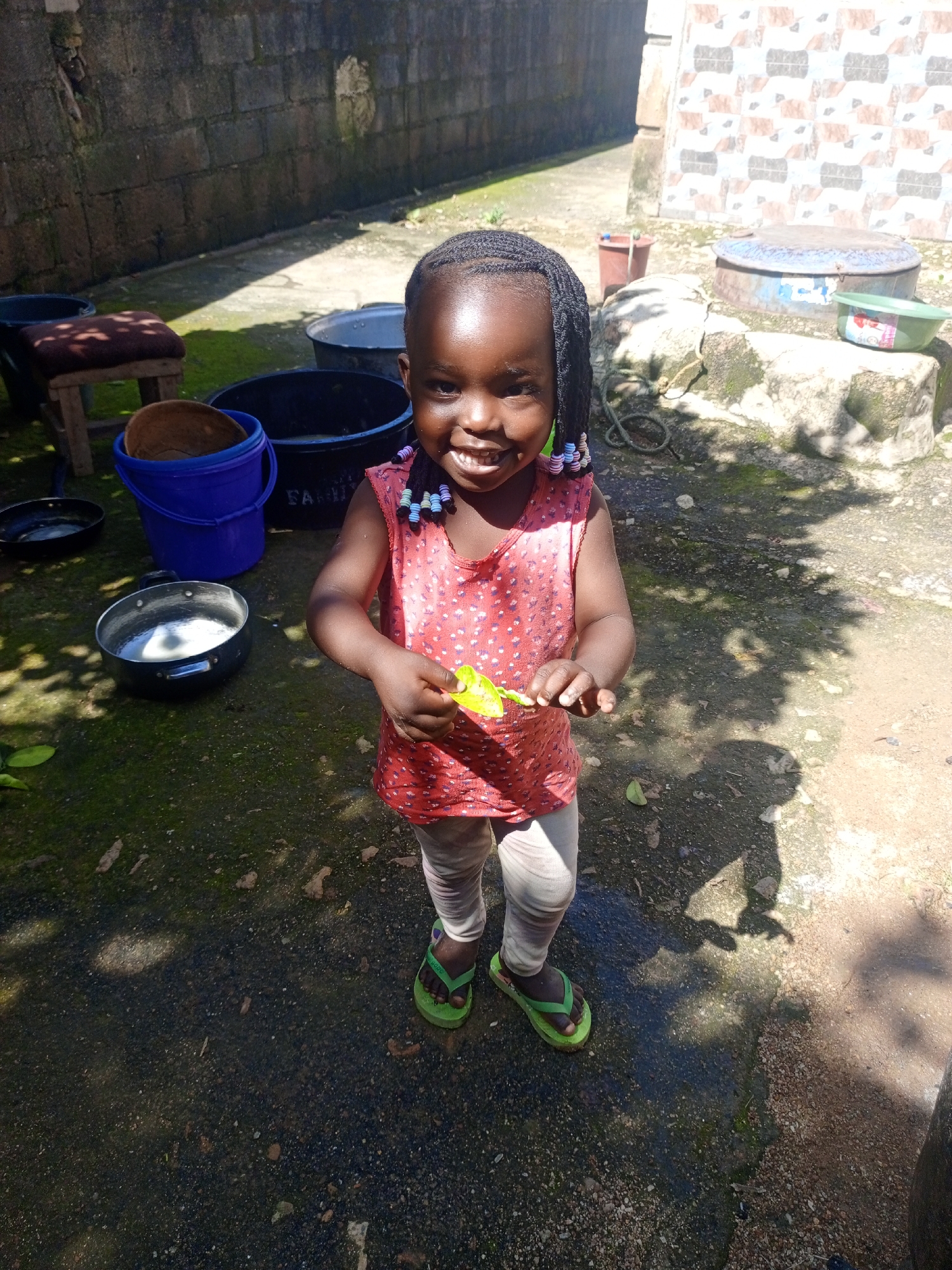 A young girl stands outdoors in what appears to be a courtyard or open area in Jos, Nigeria. She is smiling at the camera, holding a green object in her right hand. She is wearing a red sleeveless top with white polka dots, and light-colored leggings or pants. Her feet are clad in green flip-flops. Her hair is styled in braids with colorful beads.
The ground is a mixture of concrete and dirt, with some patches of moss or grass. In the background, there are several objects, including large black plastic basins, a blue bucket with a brown object inside, a smaller black pan containing a white liquid, and a larger black pan on the ground to the left. A brick wall runs along the left side of the frame, and a tiled wall is visible in the upper right background. There are also some metal containers and a wooden stool with a cushioned top. The scene is illuminated by bright sunlight, casting shadows on the ground. The overall impression is of a domestic outdoor space.