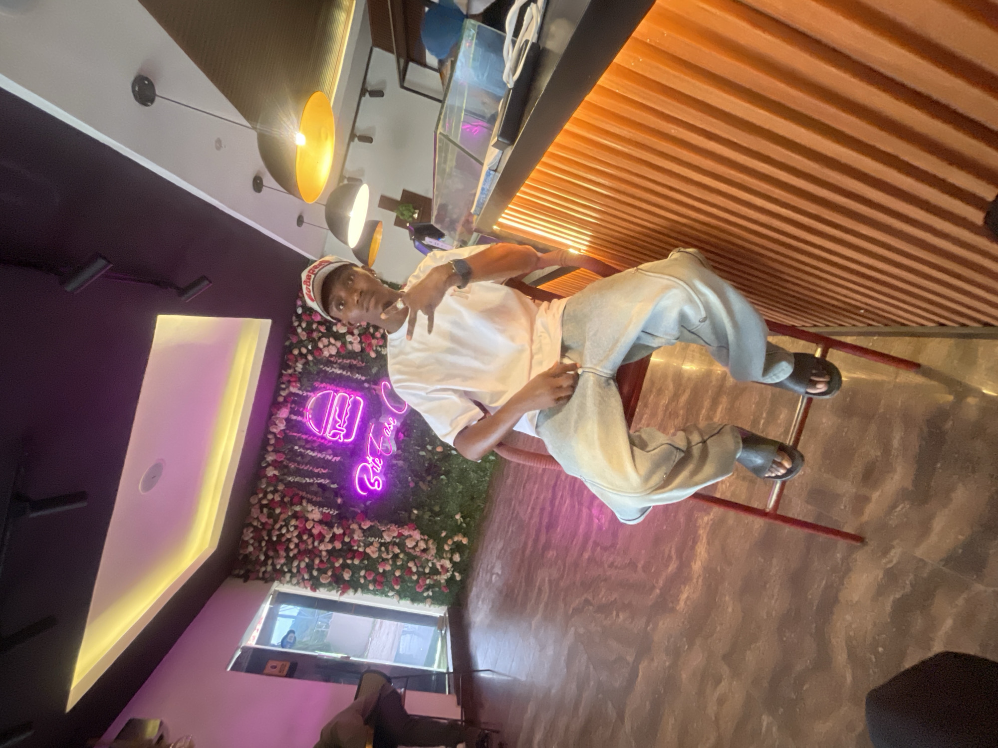 A young man is sitting on a red chair in what appears to be a restaurant or cafe, striking a pose for the camera. He is wearing a white t-shirt, light-colored pants, a cap, and slides. Behind him is a wall decorated with flowers and a neon sign that reads "Bite Case". The lighting in the cafe is a mix of natural light from a window and artificial light from hanging lamps. The cafe also features a wooden panel design on one side of the ceiling and a counter or serving area on the other side. Based on the information, the scene takes place in the city of Adankolo, Nigeria.
