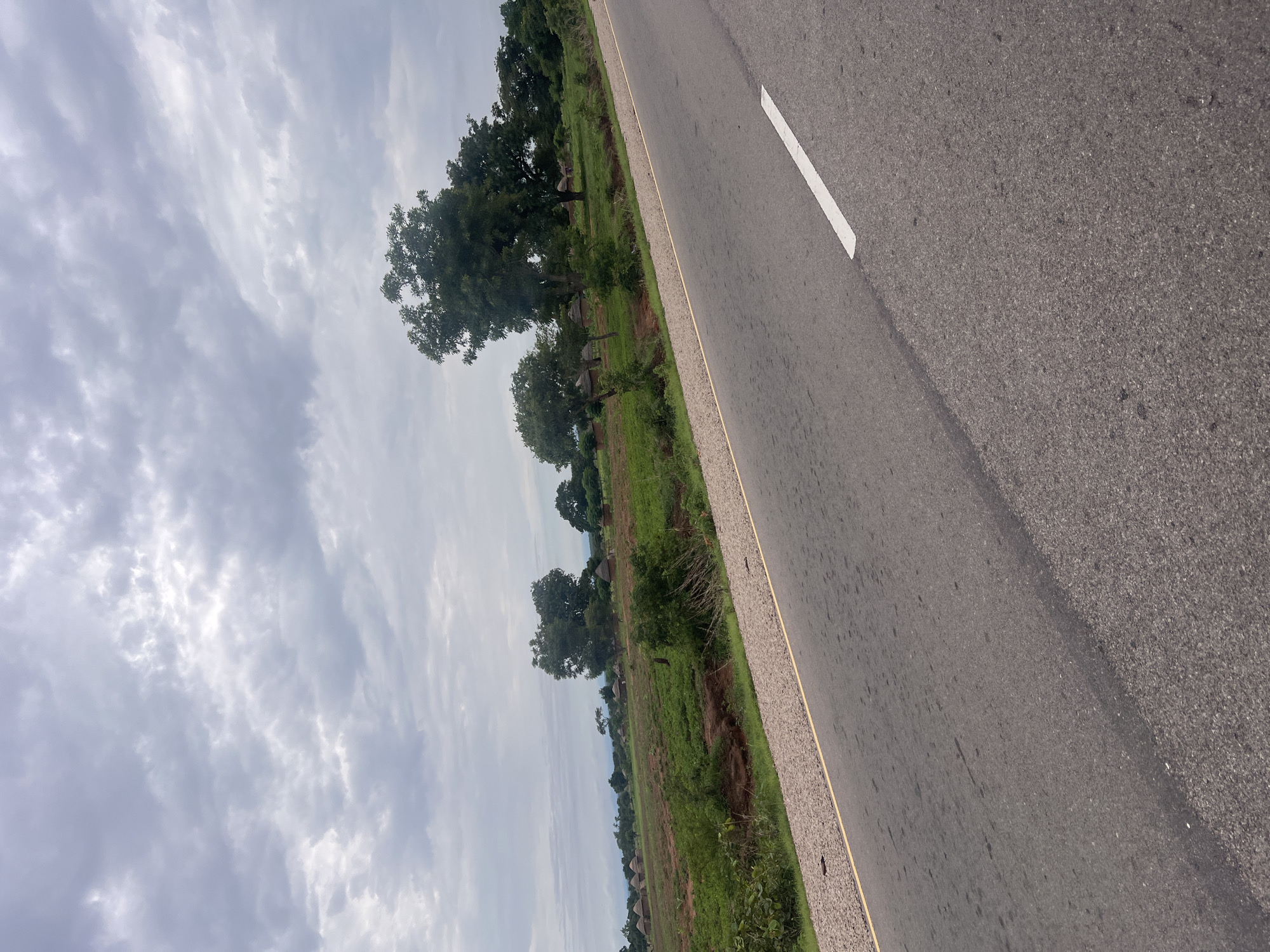 The media file depicts a road in Minda, Nigeria, with a roadside scene of vegetation and scattered huts. The road, made of asphalt, is the dominant element in the foreground, marked by a white line along its edge. To the left of the road, a strip of dry grass and soil separates it from a lush green field. This field is dotted with trees of varying sizes, and in the distance, several traditional thatched-roof huts are visible. The sky above is overcast with a heavy cloud cover, suggesting a potentially dull or rainy day. The lighting indicates it is likely daytime, but the overcast conditions limit direct sunlight. The overall scene conveys a sense of rural life in Nigeria, with the road serving as a connection to the wider world and the surrounding landscape reflecting the local environment. There are no people or explicit activities captured in the image, focusing instead on the static elements of the setting.