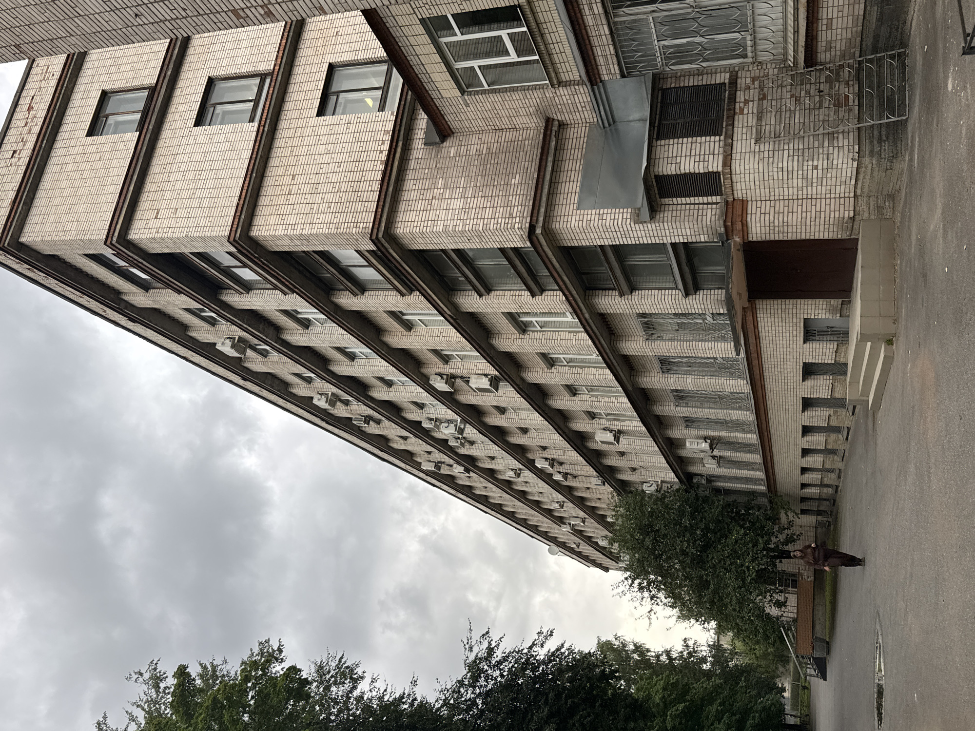 In Sinyavino, Russia, a woman in a brown outfit walks across a paved courtyard in front of a tall, multi-story brick building. The building has a light-colored brick facade and numerous windows, many equipped with air conditioning units. The courtyard is mostly bare except for a puddle of water on the pavement and some trees to the left and in front of the building. The sky is overcast and gray. The building appears to be an institutional or residential structure.