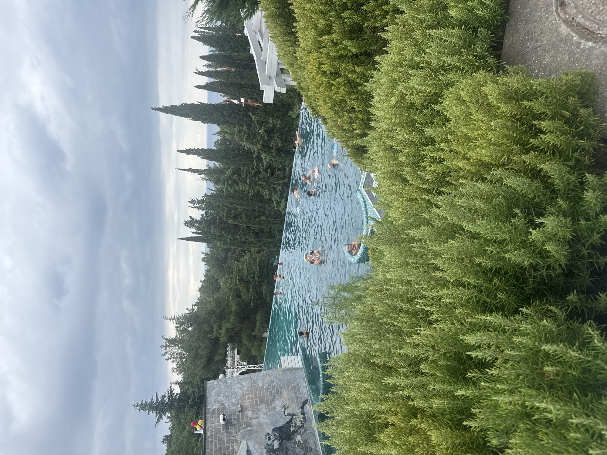 The image shows an infinity pool with several people swimming and relaxing. The pool is surrounded by lush green vegetation and tall cypress trees. In the background, the sea is visible under a cloudy sky. The pool is adjacent to a building with a white roof and decorative white architectural elements. On the left side of the pool, a grey brick wall features a mural depicting a person with a camera. On top of the wall, a person in a yellow hat and red jacket stands near a speaker. The overall scene suggests a resort or recreational area in Yalta, Ukraine.
