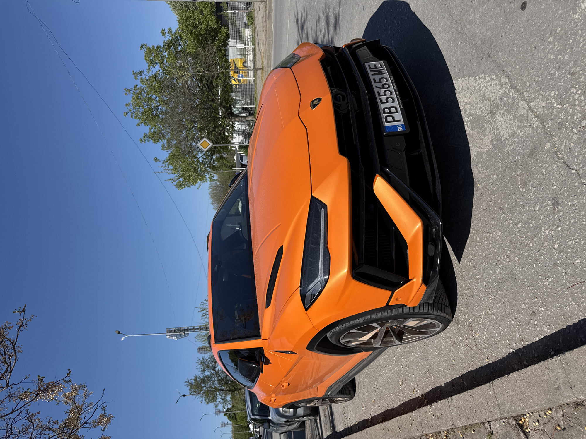 A Lamborghini Urus is parked on the side of a street. The car is painted orange with black accents. The vehicle is parked on an asphalt surface, likely a road. A streetlight and trees are visible in the background. The license plate reads "PB 5565 ME" and has a "BG" country code, indicating Bulgaria. The photo was likely taken in Plovdiv, Bulgaria.