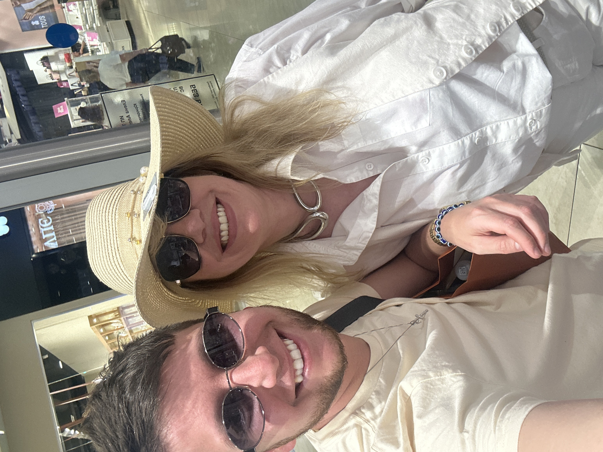 A selfie taken in an indoor commercial environment, possibly a shopping mall, shows two individuals. The person on the left, a male with short brown hair and a beard, wears sunglasses and a light-colored t-shirt with a silver necklace. The person on the right, a female wearing a wide-brimmed straw hat with pearl embellishments and sunglasses, has blonde hair and a bright white collared shirt. She is wearing a chunky silver necklace and a blue and gold beaded bracelet. A brown bag is visible between them. In the background, a store with displays of merchandise and other shoppers can be seen. The text on a sign in the background appears to be in Russian, and one store prominently displays the brand "TOUS". Based on the presence of Russian text and the "TOUS" brand, which has numerous locations in Russia, this scene is likely in Moscow, Russia.