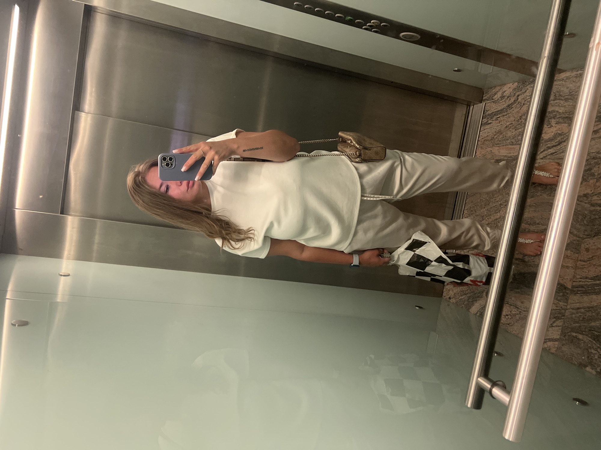 A young woman is captured in a full-body selfie taken in a reflective elevator interior in Budva, Montenegro. She stands facing the reflective surface, holding a grey smartphone with a triple-lens camera in her right hand to take the picture, partially obscuring her face. Her long, light brown hair falls over her shoulders.
She is dressed in a matching light-colored outfit, consisting of a short-sleeved top and trousers, possibly white or a very light beige/grey. Accessories include a small, light brown patterned crossbody bag with a gold chain strap, likely a Guess brand. On her left wrist, she wears a light-colored smart watch, and a ring is visible on her right hand. A tattoo of Roman numerals, "IIV.VMMXVII", is visible on her left forearm. Her right foot is adorned with sparkling, jeweled sandals or foot jewelry. In her left hand, she holds a black and white checkered bag with some red detailing.
The elevator features polished metal walls and ceiling panels, a row of elevator buttons at the top, and a rich, veined granite or marble floor. Metal handrails are visible on the right side, and a semi-transparent or frosted glass panel is on the left. The scene is well-lit by interior artificial lighting.