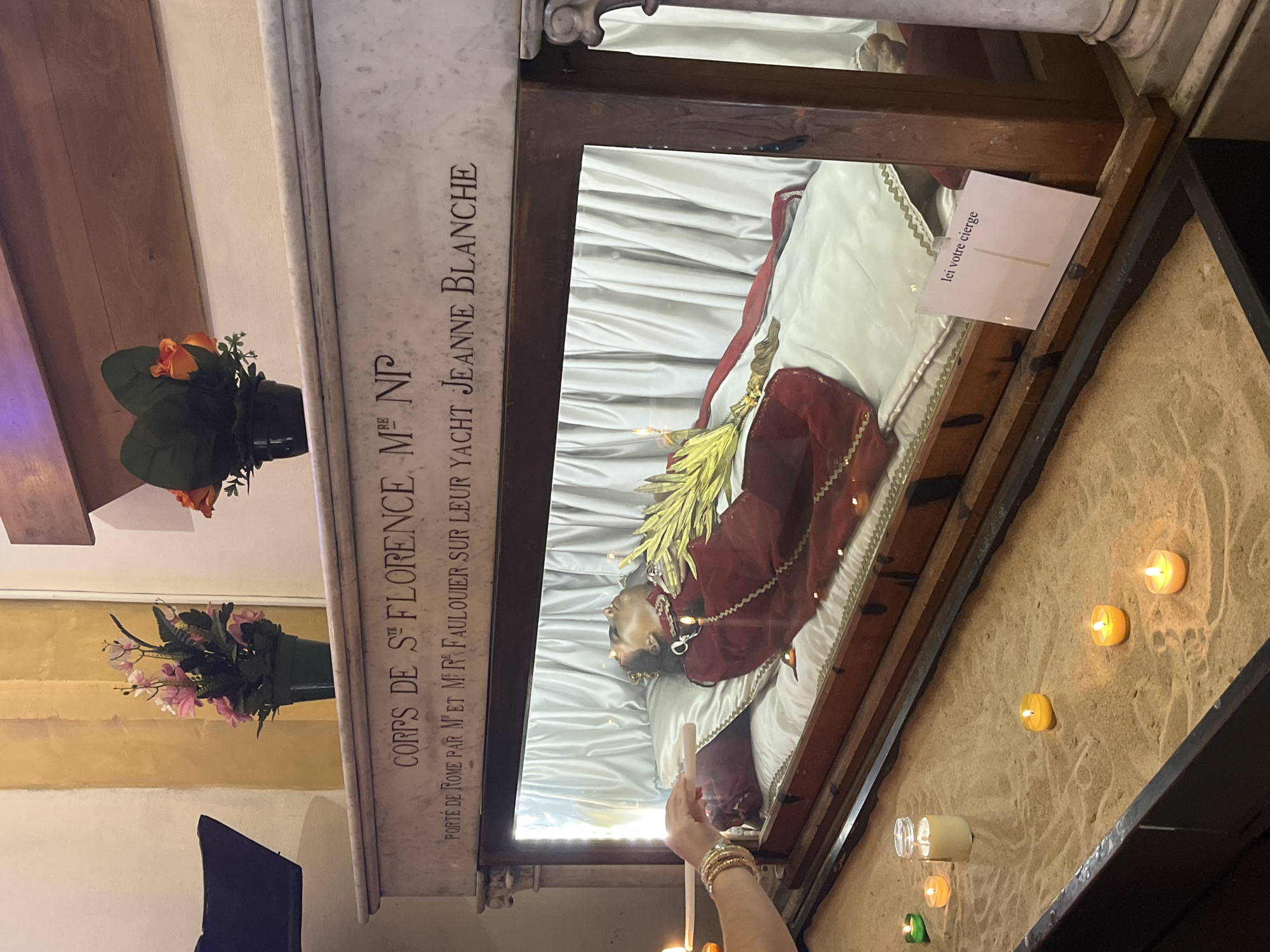 The image depicts the reliquary of Saint Florence in the Church of Saint-Pierre in Palavas-les-Flots, France. The reliquary, a glass-covered sarcophagus, displays the preserved body of the saint adorned in red robes and a crown. The body rests on a white satin cushion, with a golden scepter-like object beside it. The sarcophagus is placed on a marble base inscribed with text, including "CORPS DE STE FLORENCE" and "PORTE DE ROME PAR M ET MME FAULOUIER SUR LEUR YACHT JEANNE BLANCHE". In front of the reliquary, a sand-filled area contains several lit votive candles of various sizes and colors, and a jar with a candle. A person's hand, adorned with bracelets, is reaching towards the reliquary, holding a thin object, possibly to light a candle. In the background, floral arrangements are visible on top of the reliquary, and a piece of text that reads "Ici votre cierge" is placed on the edge of the sarcophagus. The overall environment suggests a religious setting, possibly a church or chapel, with soft lighting and a somber atmosphere.