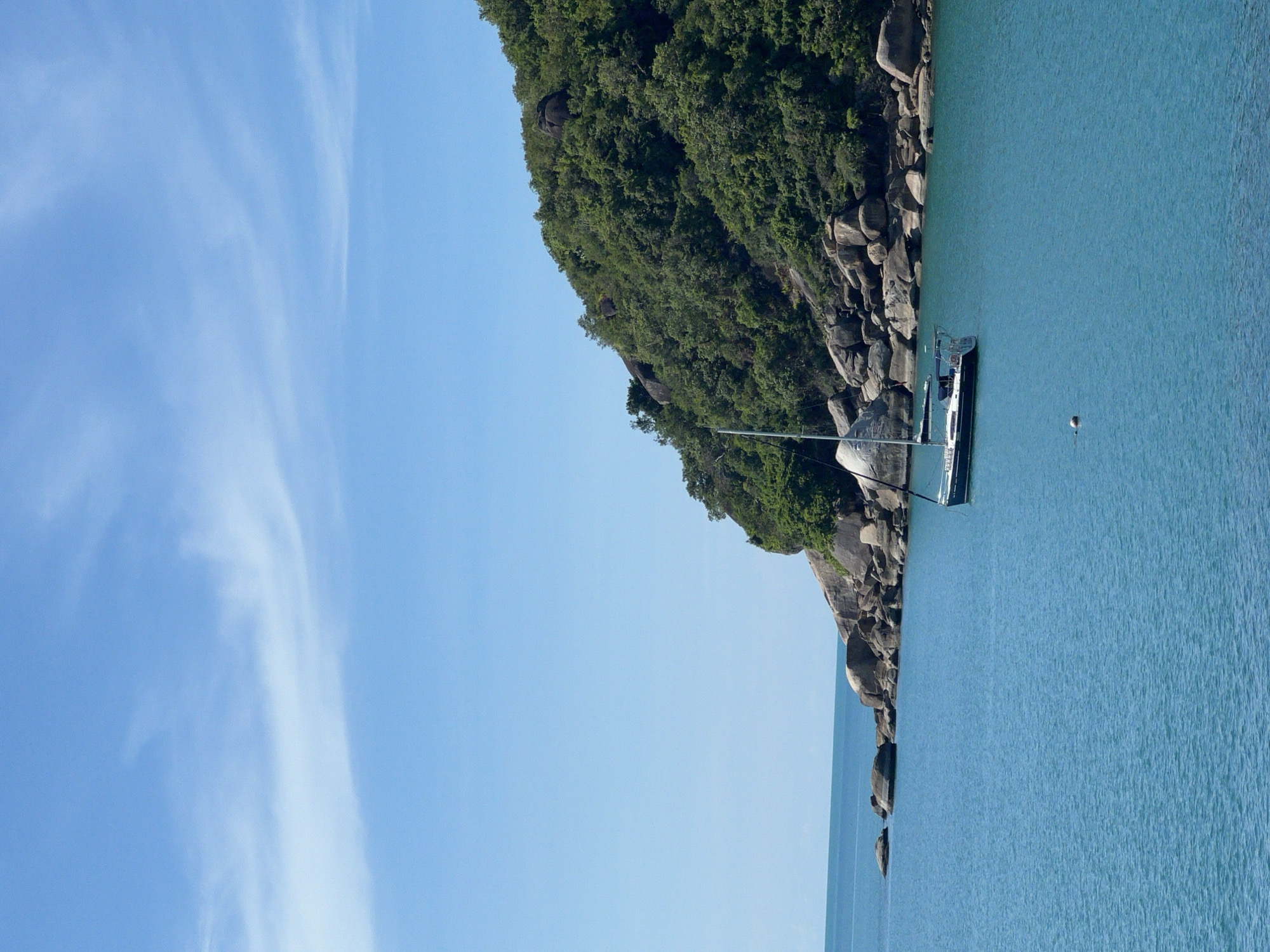 The image depicts a calm, clear blue ocean with a sailboat anchored in the foreground. The sailboat is dark in color with a white sail. Behind the sailboat, a rocky, tree-covered island slopes upwards towards a bright blue sky with thin white clouds. The coastline features large, rounded boulders interspersed with lush green vegetation. The location is Baan Chaweng Noi, Thailand.
