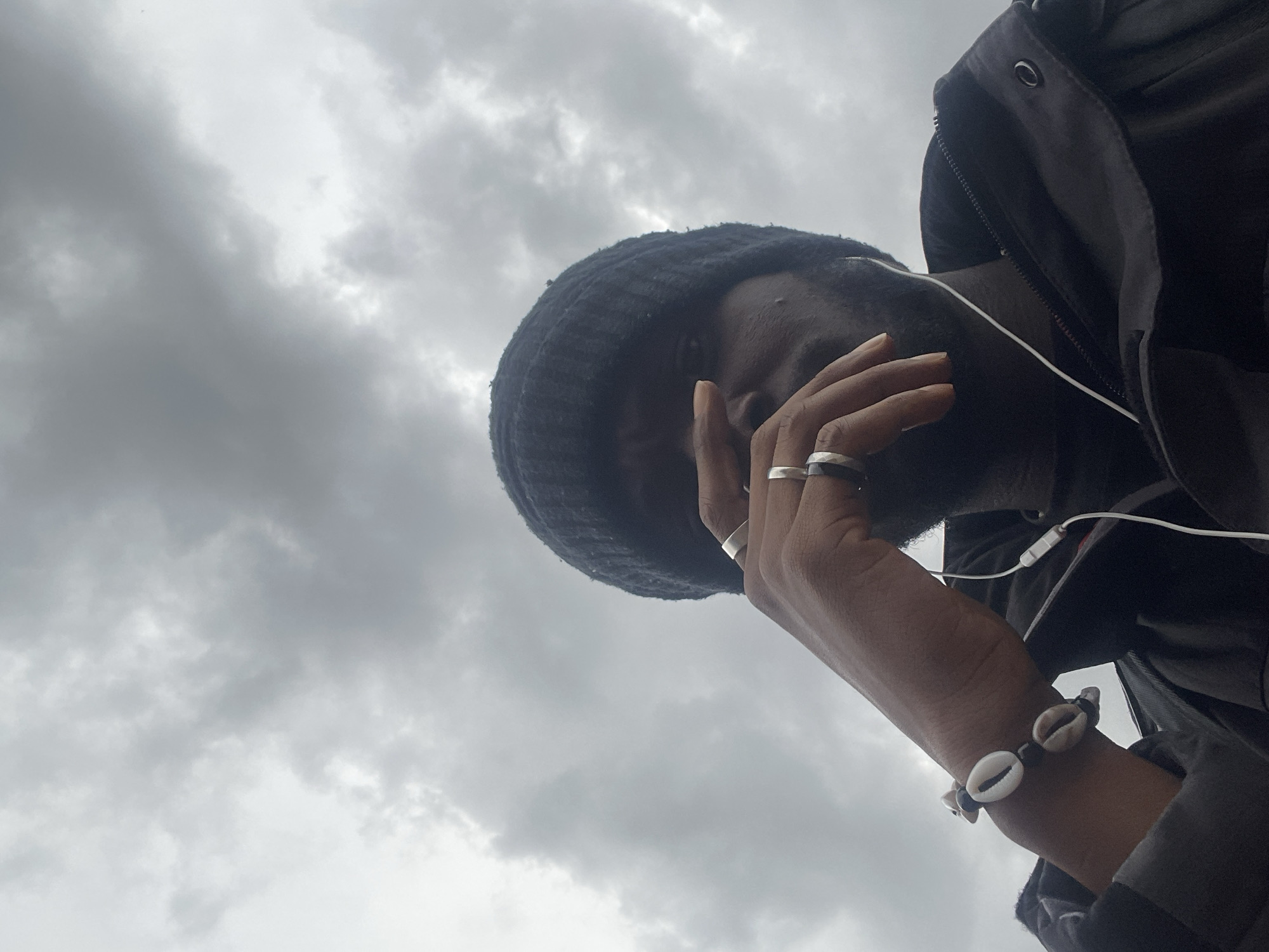 A low-angle shot captures a person with dark skin against a dramatically overcast sky. The subject is seen from below, looking downward or to the side, with their right hand raised to partially cover their face. They wear a dark, possibly knitted, hat or beanie and a dark jacket, featuring a visible zipper and a metallic snap button near the shoulder. White wired earphones are draped around their neck and near their ear. The person’s right hand is adorned with multiple silver-toned rings on their fingers, and a bracelet crafted from cowrie shells and dark beads on their wrist. The background is dominated by thick, grey and white clouds, indicating a heavily overcast daytime sky. The pose suggests a moment of contemplation or a stylized shot. No text is visible in the media. The implied setting is Jos, Nigeria.