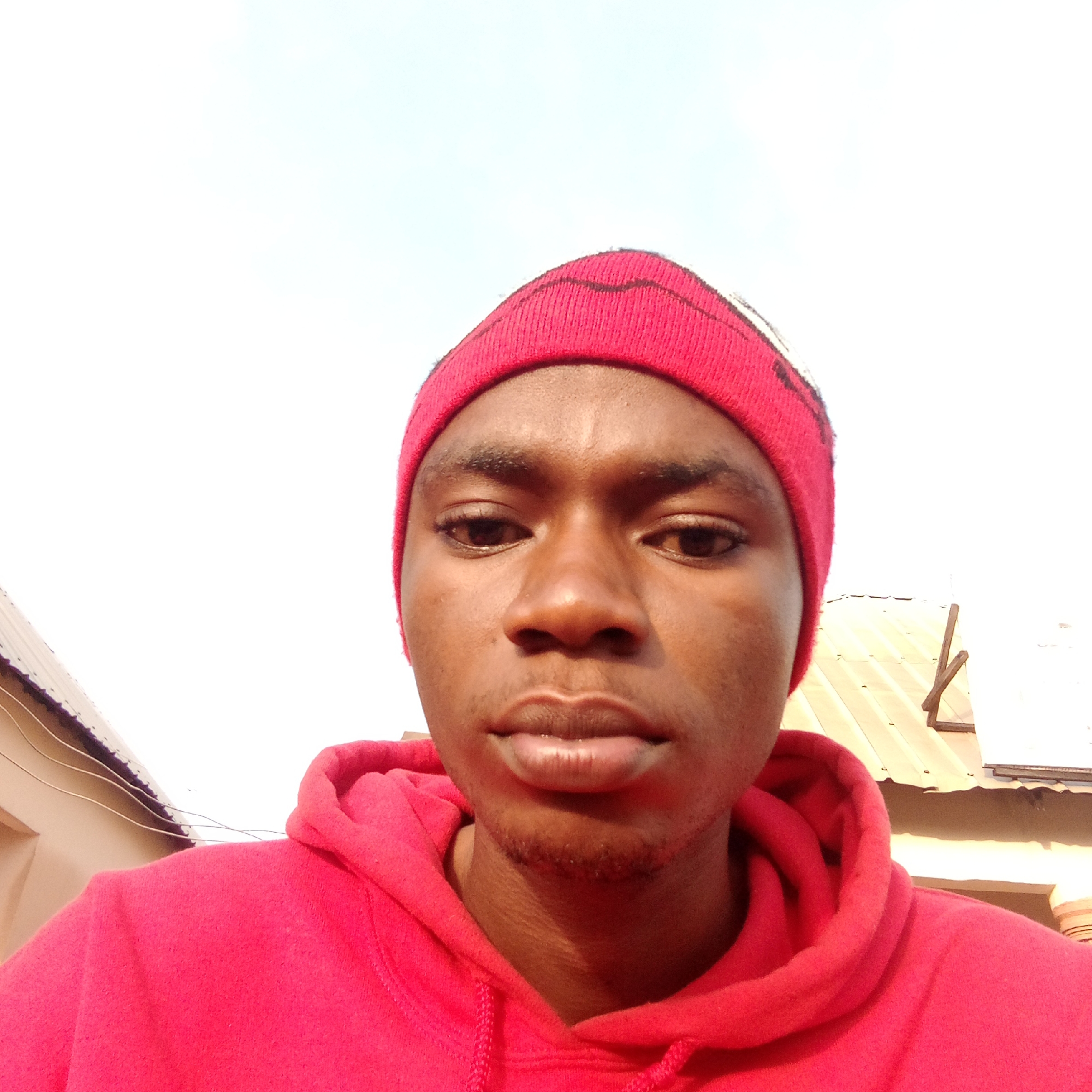 A close-up selfie of a young man wearing a red beanie and hoodie. The background features a building and a bright sky, suggesting an outdoor setting. Based on the prompt, this location would be assumed to be in Mararaba, Nigeria.