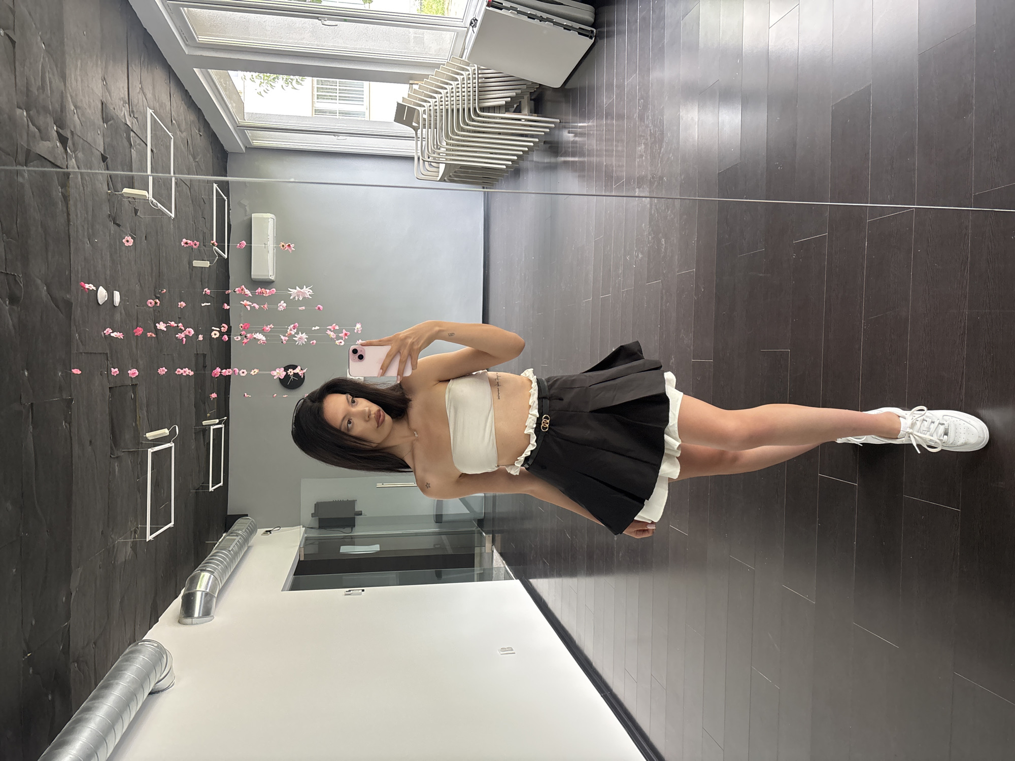 A young woman with dark hair, wearing a white bandeau top and a black pleated skirt with white ruffles, poses for a mirror selfie in a studio or dance space. She is holding a pink smartphone in her right hand. The room has dark wood floors, grey walls, and a black ceiling with exposed silver ventilation pipes. Decorative pink and white paper flowers hang from the ceiling. In the reflection of the mirror, a row of stacked chairs and folded tables are visible near a large window. The location is a studio space in the city of Sofia, Bulgaria.