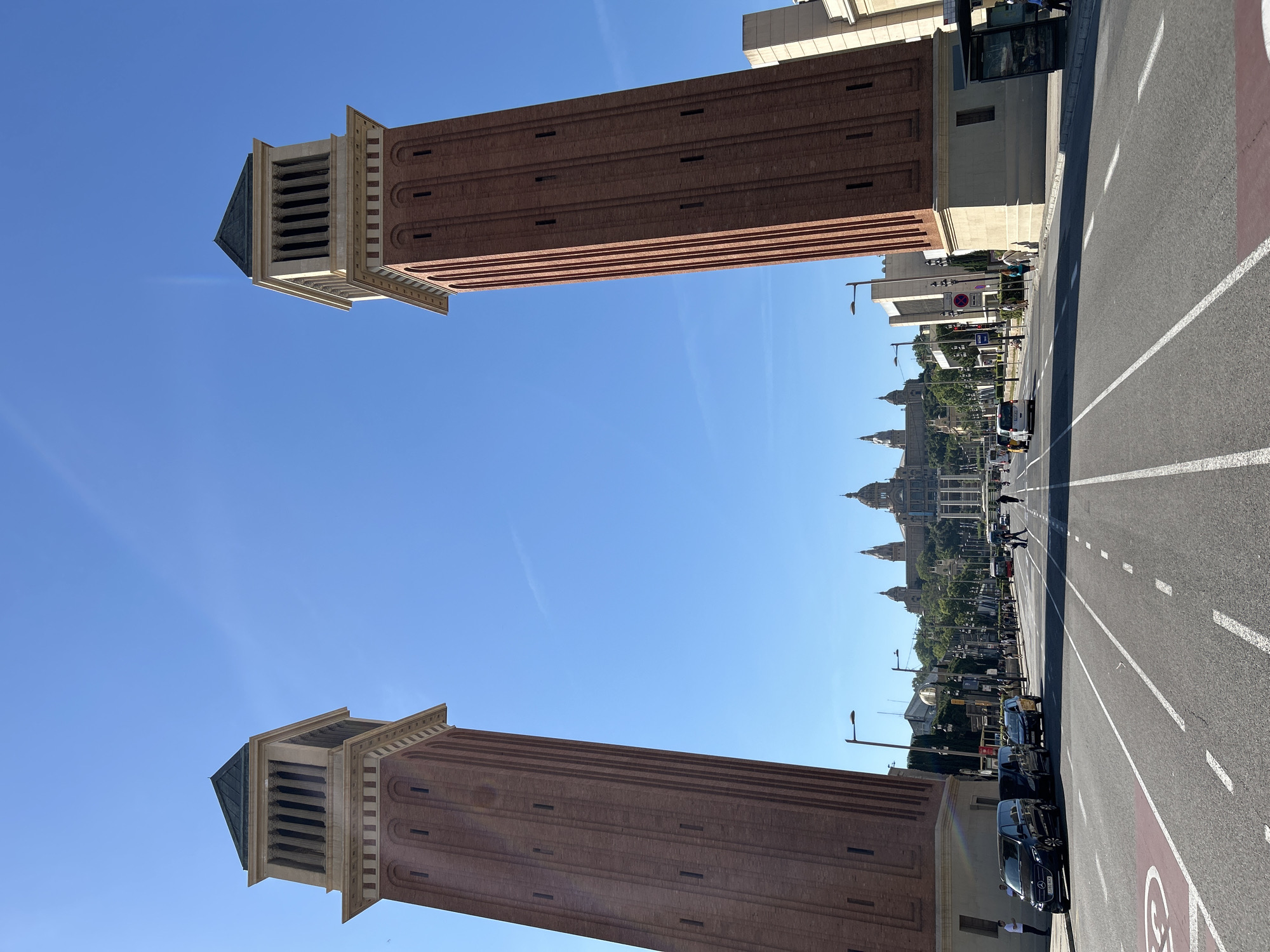 The image captures a view along Avinguda de la Reina Maria Cristina in Barcelona, Spain, looking towards the Palau Nacional. Two distinctive Venetian Towers flank the avenue. The structures are made of reddish-brown brick with cream-colored accents at the top. A blue sky is visible between the towers. The avenue shows road markings and traffic, including cars and pedestrians. In the distance, the Palau Nacional, a large building with domes, stands at the end of the avenue.
