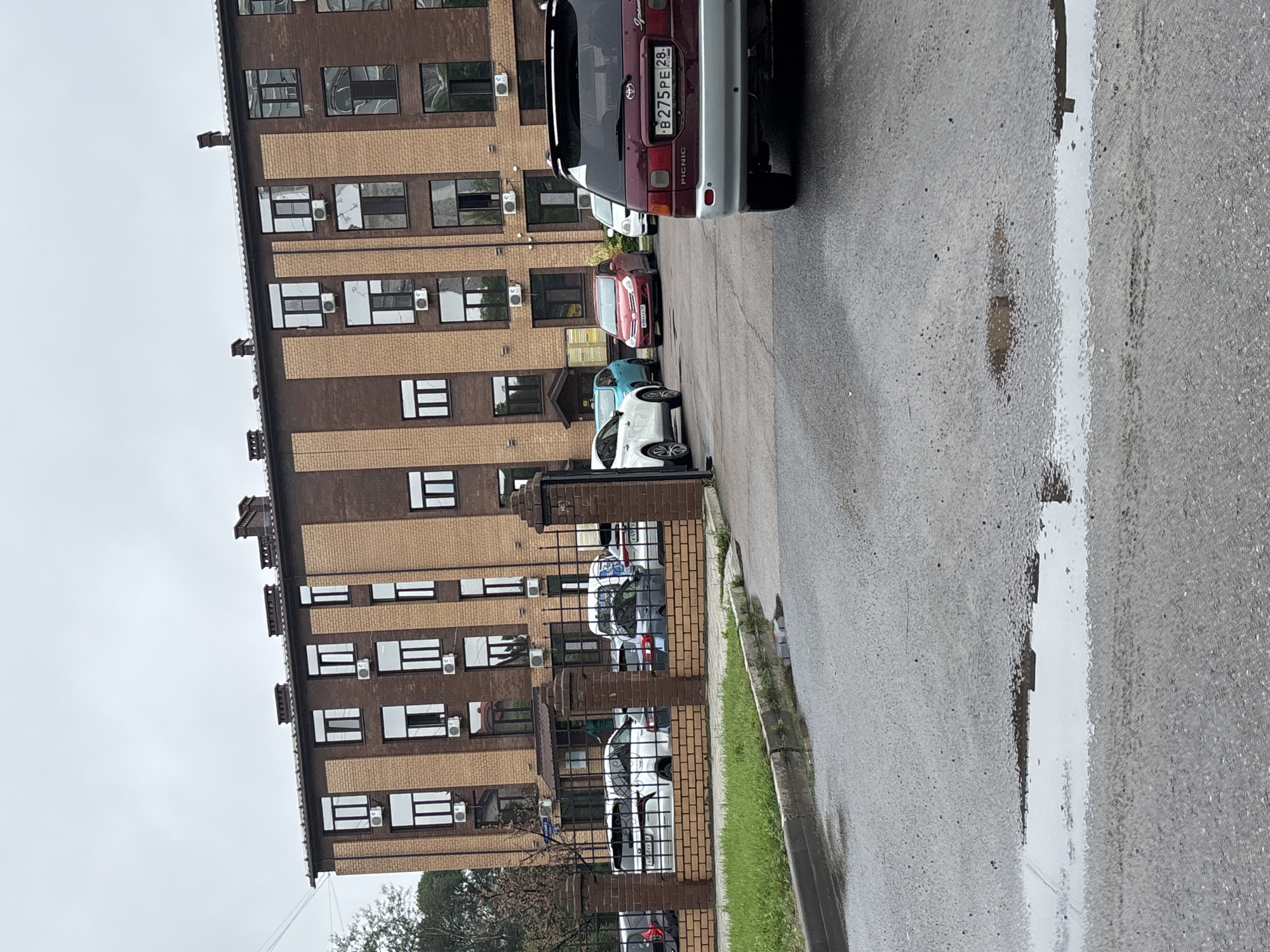The image captures a street scene in Blagoveshchensk, Russia, on a cloudy day. A multi-story brick building with numerous windows and air conditioning units dominates the background. In front of the building, a black metal fence encloses a parking area where several cars are parked, including a red Toyota Picnic with a license plate reading "B275PE 28". To the left of the fence, a strip of green grass runs along a curb, and further left, a tree is partially visible. The foreground shows a wet asphalt road with puddles reflecting the overcast sky. There are no people clearly visible in the image. The overall environment suggests a residential or commercial area with limited pedestrian activity at the time of the photograph.