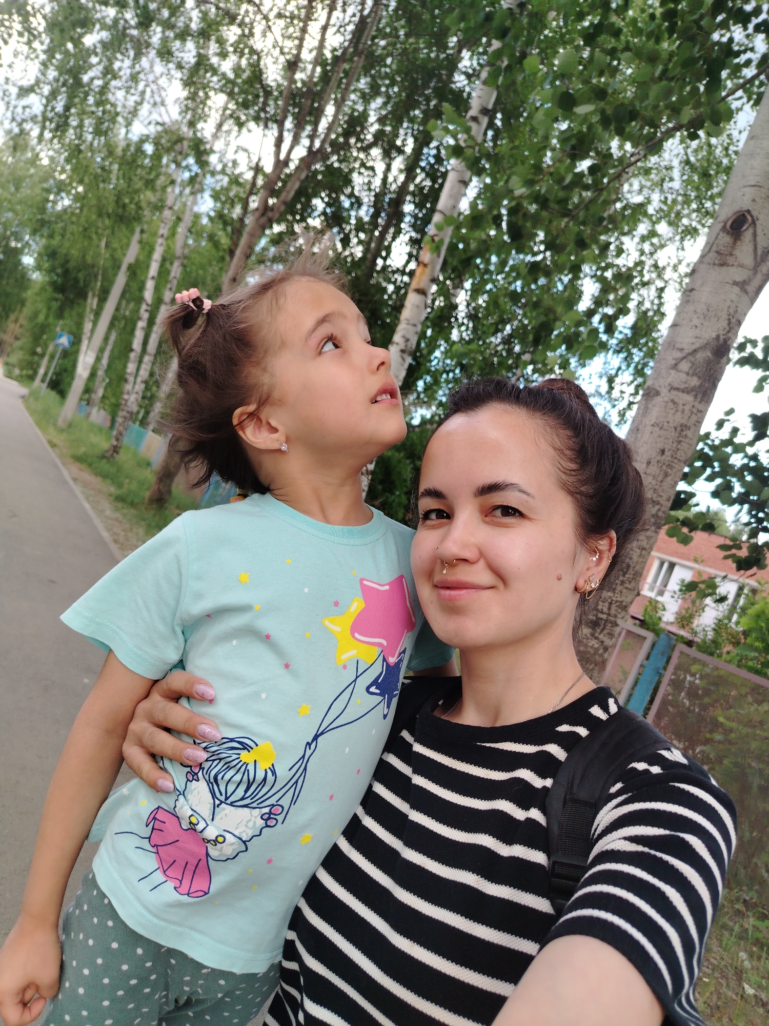 The image captures a woman and a young girl, possibly a mother and daughter, standing in an urban environment.
**People:** The woman has dark hair pulled back in a bun, fair skin, and several piercings in her ear and nose. She wears a black and white striped shirt with a black backpack. The girl is younger with light brown hair partly tied back, fair skin, and wears a light blue t-shirt with a cartoon design.
**Actions and Interactions:** The woman is holding the camera in a selfie position while the girl is looking upwards with interest.
**Objects:** A road and trees line the foreground. A metal and wooden fence with a building in the background are also visible.
**Environment:** The location is in the city of Noviy, Russia. The weather appears pleasant, with abundant greenery suggesting either spring or summer. The scene suggests an urban public space, possibly a park, neighborhood, or street with residential areas nearby.