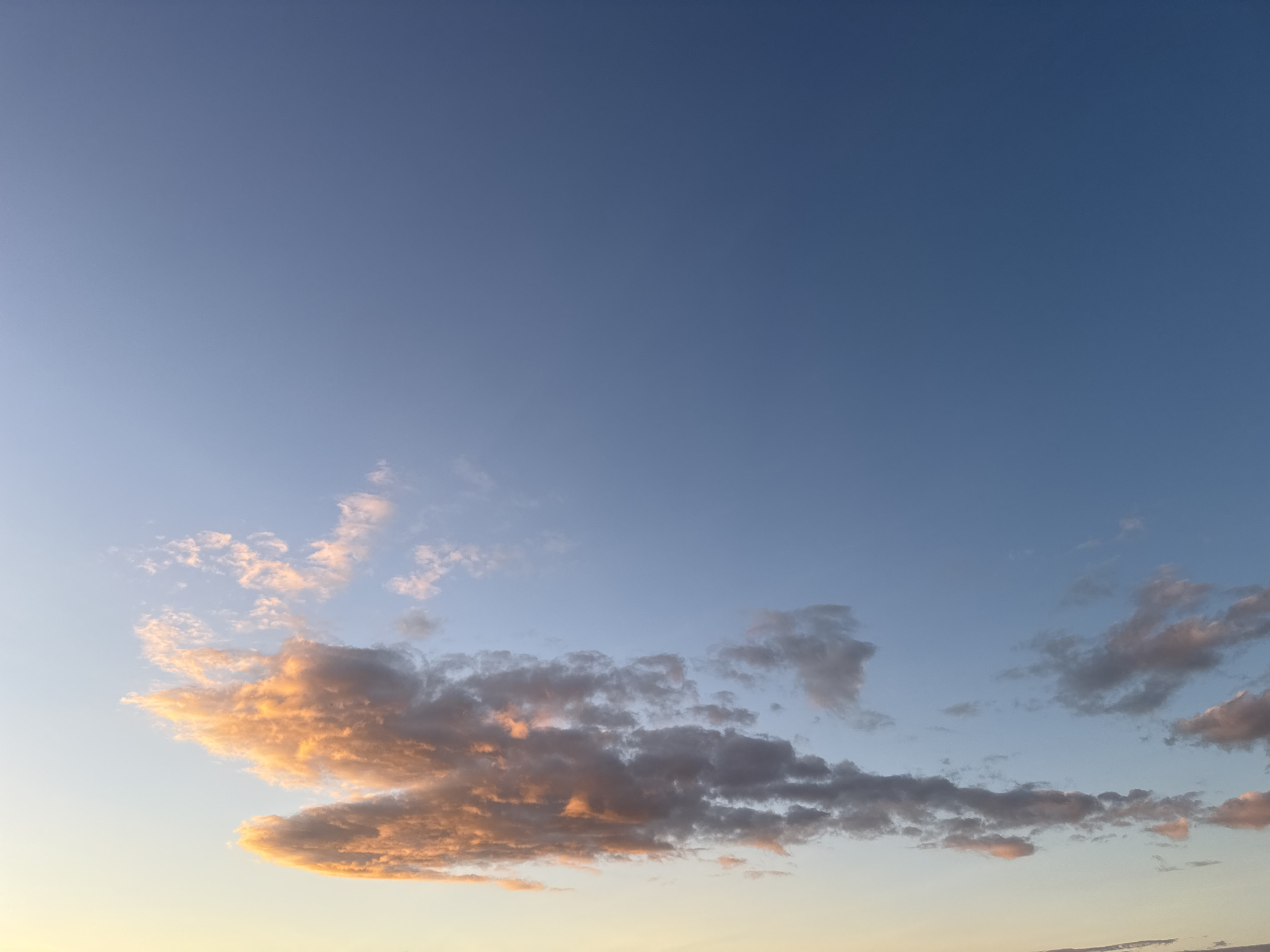 The image shows a clear blue sky with scattered clouds illuminated by warm, orange light, suggesting either sunrise or sunset. The clouds are of varying densities, with some appearing thin and wispy, while others are more substantial and textured. The lower part of the image shows a faint transition to a lighter, yellowish hue near the horizon, indicative of the low angle of the sun. No people, objects, or specific landmarks are visible, indicating an open sky view, likely from an outdoor location. The image composition focuses entirely on the sky and cloud formations. The precise location within Бараўлянскі сельскі Савет, Belarus, cannot be determined from the visual information provided in the image.