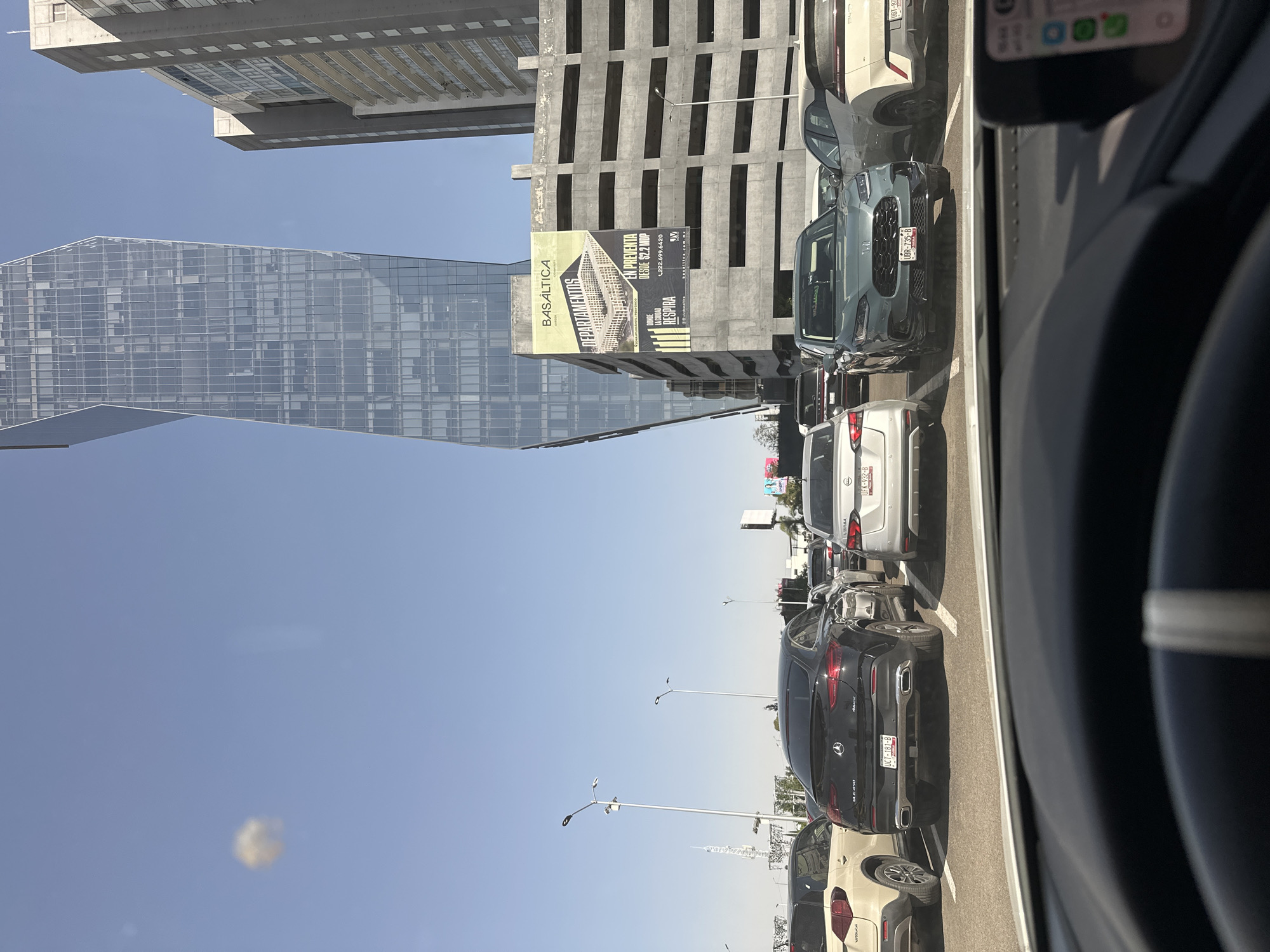 An urban street scene unfolds from the perspective of a vehicle, revealing a line of cars and prominent buildings under a clear, bright blue sky. The dashboard and part of a steering wheel are visible on the right, indicating the viewpoint is from inside a car. Several cars are stopped or in slow-moving traffic ahead; a dark SUV displays license plate UBR-735-B, a white SUV has UFK-932-B, and a black Mercedes-Benz GLE 350 bears UCT-181-B.
On the left, a striking, modern skyscraper with a reflective glass facade rises against the sky. Adjacent to it, a multi-story concrete building, possibly under construction or a parking structure, features a large banner. This banner advertises "BASÁLTICA DEPARTAMENTOS" (apartments) for "EN PREVENTA" (pre-sale) "DESDE $2.2 MOP" (from $2.2 million Mexican Pesos). The slogan "DONDE LA CIUDAD RESPIRA" (Where the city breathes), a phone number "222.699.6420," and the website "basaltica.mx" are also clearly visible. The overall scene suggests a sunny, daytime setting in a bustling city, likely in Mexico, given the Spanish language and currency reference.
