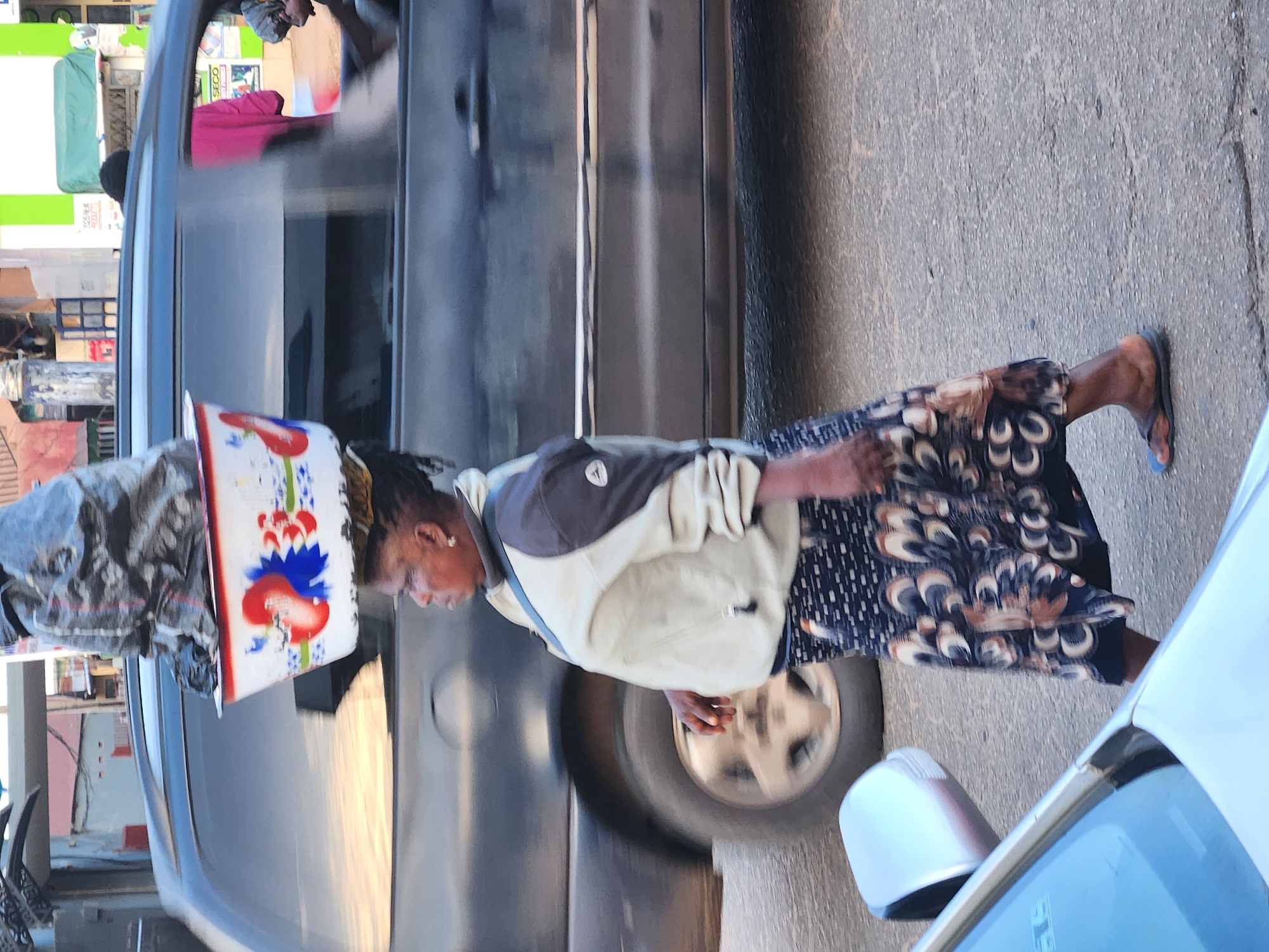 A woman walks along a street, balancing a stacked load of items on her head, near vehicles in the city of Unknown, Unknown. The woman is wearing a light-colored jacket with dark gray accents and a patterned skirt in shades of brown, white, and black. She also wears a pair of blue flip-flops on her feet. On her head, she supports a stack of items including a red, white, and blue bucket, and covered with a gray, patterned cloth. A black SUV is parked alongside the woman, with the reflections visible on the car's windows. To the right of the woman, a white vehicle is partially visible, including its side mirror and front part of the windshield. The street appears to be paved and the overall scene suggests an urban setting.