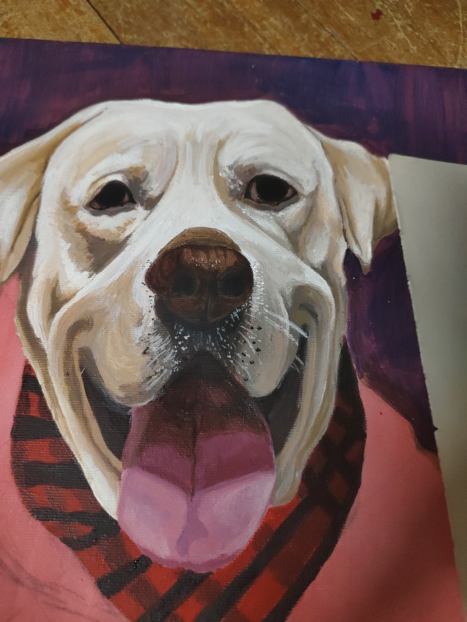 A painted portrait of a light-colored dog, likely a Labrador Retriever, is depicted in a close-up, front-facing view, observed within an indoor environment located in Davao City, Philippines. The artwork, rendered in what appears to be oil or acrylic, showcases the dog with a wide, open-mouthed expression and its pink tongue extended. The dog's fur is predominantly cream-colored with subtle beige and brown shading, featuring prominent dark eyes and a textured dark brown nose. A red and black plaid bandana adorns its neck. The painted background consists of a dark purple upper section transitioning to a solid coral-pink lower section. Visible brushstrokes and canvas texture are present across the painting. The artwork is positioned such that a section of a wooden floor is visible above its top edge, indicating a physical setting where the painting rests or is displayed.