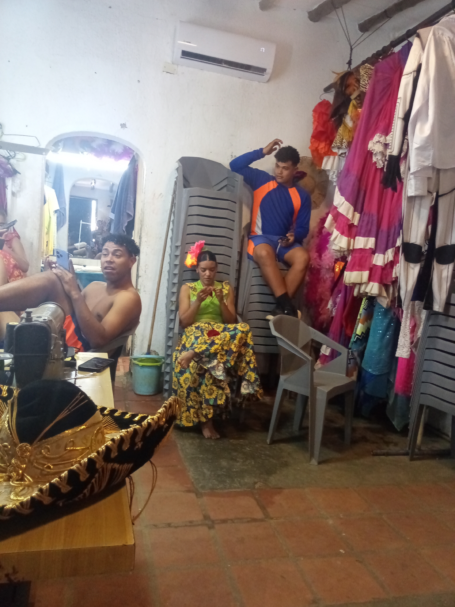 This image captures an indoor scene, likely a costume preparation area or backstage room, in Porlamar, Venezuela. The setting features white walls, a tiled floor, and an arched doorway leading to another room where more people are visible. An air conditioner unit is mounted high on the wall.
Three main individuals are present. On the left, a shirtless man with dark, curly hair sits, holding a smartphone and looking towards the viewer with a slightly surprised expression. He is positioned near a wooden table that holds a sewing machine and a prominent, ornate black and gold sombrero. In the center, a woman with a flower in her dark hair is seated on a low chair. She wears a green top and a long, vibrant yellow skirt with a floral pattern and appears to be focused on something she holds in her hands, possibly engaging in a crafting or sewing activity. A small blue bucket is beside her. To the right, a young man wearing a blue and orange long-sleeved shirt and blue shorts is seated on a stack of grey plastic chairs, casually touching his hair.
The background is dominated by stacks of grey plastic chairs and a rack laden with numerous colorful costumes and garments, including ruffled dresses in shades of pink and purple, white shirts, and various other festive attire. The presence of costumes, a sewing machine, and the general atmosphere suggests a space dedicated to preparing for performances or cultural events. The individuals appear relaxed, either resting or engaged in light activity. No visible text is present in the image.