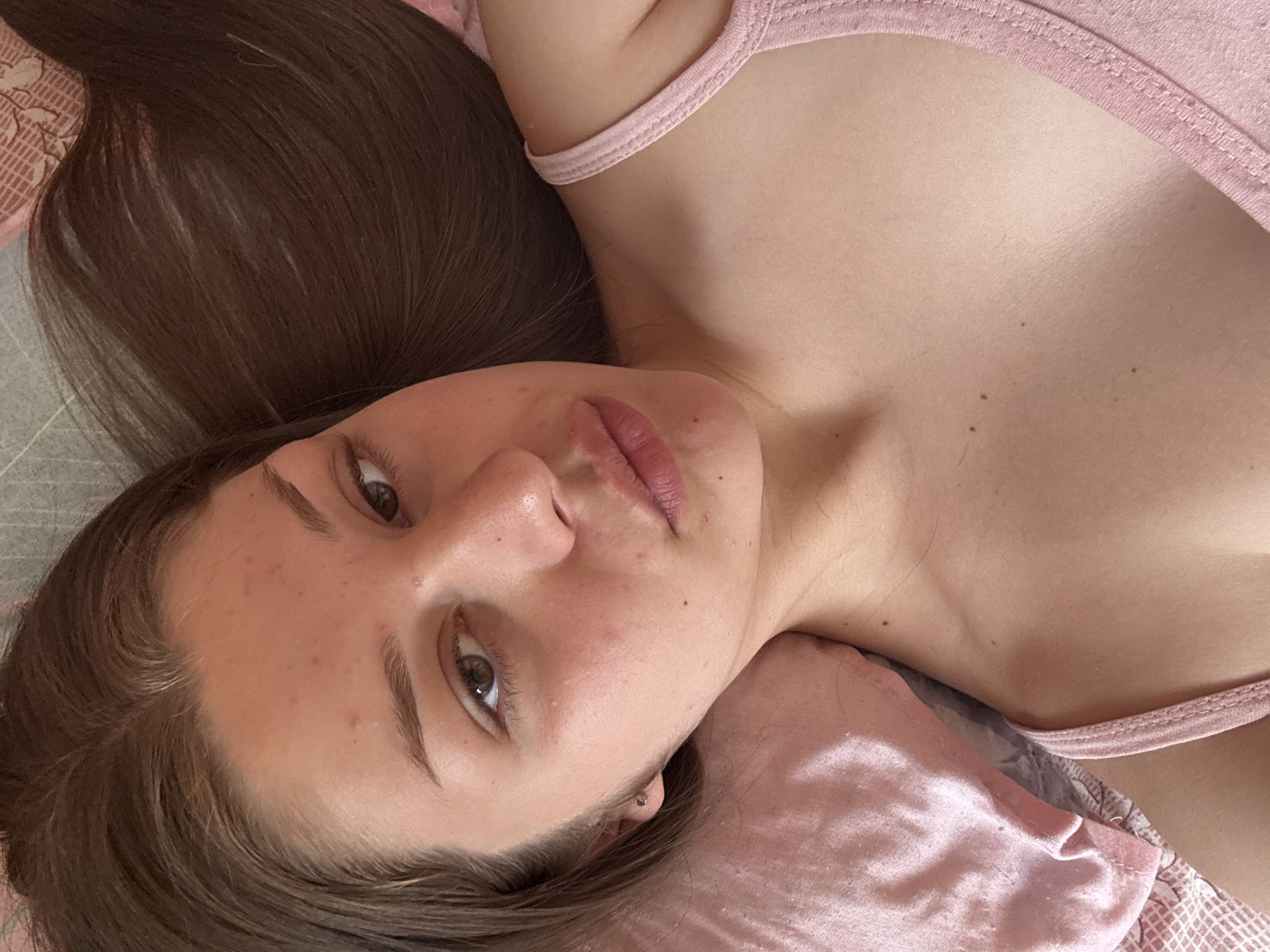 The media file is a close-up, high-angle, eye-level shot of a young person lying on their side on a bed. The person has long, dark brown hair that is spread out around them. They are wearing a light pink tank top. Their face is turned towards the camera, and their eyes are open. They have light brown eyes and dark eyebrows. Their lips are slightly parted, and they appear to be wearing some lip gloss. There are a few blemishes visible on their skin. The bedding appears to be pink and patterned. The scene is intimate and relaxed, with the subject appearing to be comfortable and at ease. The lighting suggests it could be daytime, possibly in a bedroom. There is no visible text in the image.