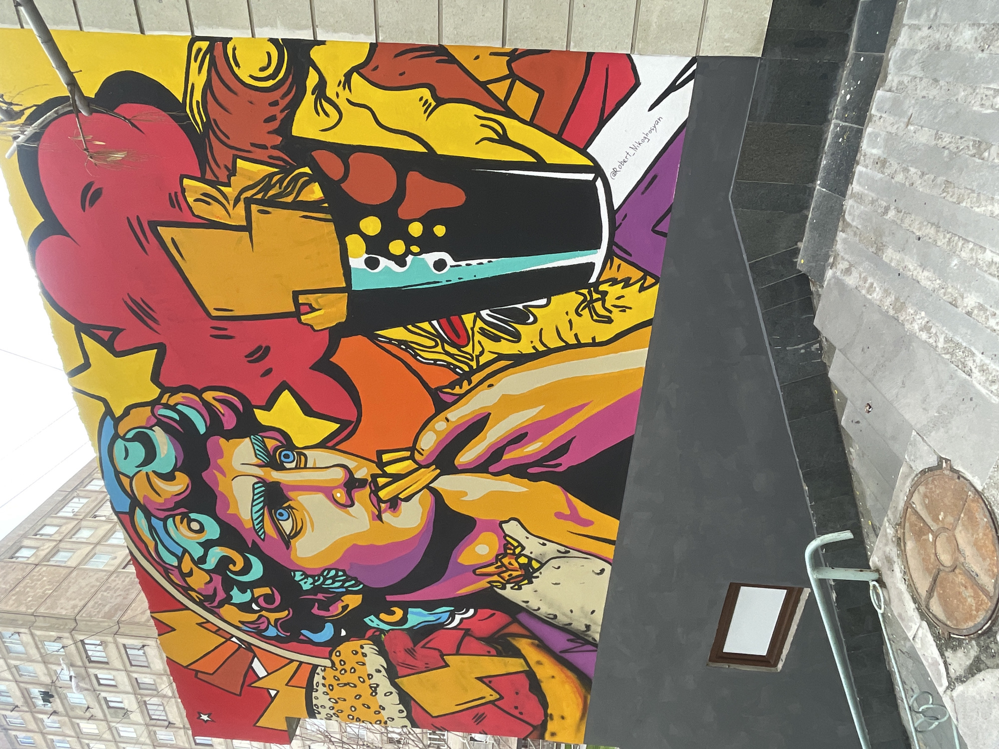 A vibrant mural adorns a wall in Yerevan, Armenia. The artwork features a stylized rendition of a classical figure, possibly David, rendered in bright, contrasting colors. He's depicted eating French fries, with a fast-food cup visible above him. The mural is filled with other food-related imagery and is signed "Robert Nikoghosyan." The wall is adjacent to a staircase with a handrail, a framed blank sign, and a manhole cover in the foreground. A multi-story building is visible in the background.