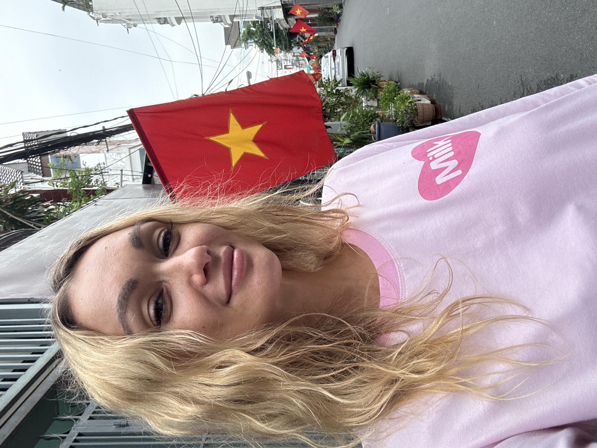 A woman with blonde, wavy hair wearing a pink sweatshirt with a heart-shaped logo containing the word "Milk" is in Đồng Nai, Vietnam. Several Vietnamese flags are visible along the street, which also includes buildings and parked vehicles. Overhead, numerous power lines crisscross.