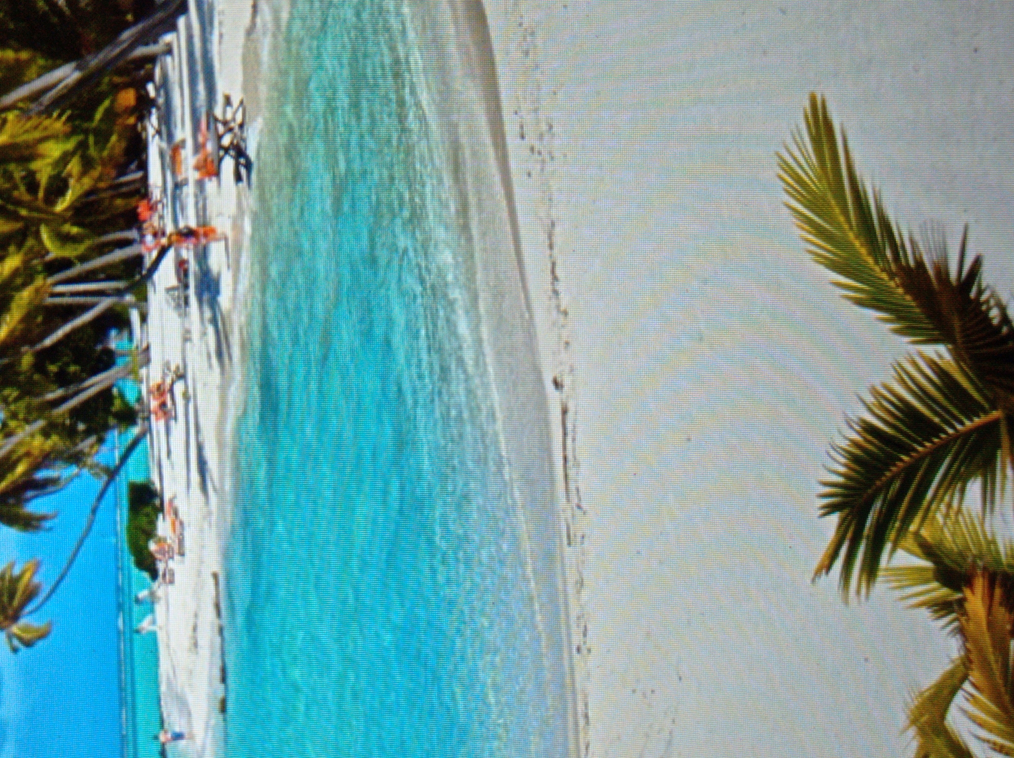 The image depicts a tropical beach scene. The foreground features a sandy beach with a few palm fronds visible at the bottom. The middle ground shows clear turquoise water with gentle waves lapping at the shore. In the background, there is a white sandy beach dotted with lounge chairs and a few people. Palm trees line the back of the beach, casting shadows. The sky is a bright blue. The location is not in the city of Mukachevo, Ukraine, as Mukachevo is a landlocked city in Western Ukraine and does not have a tropical beach.