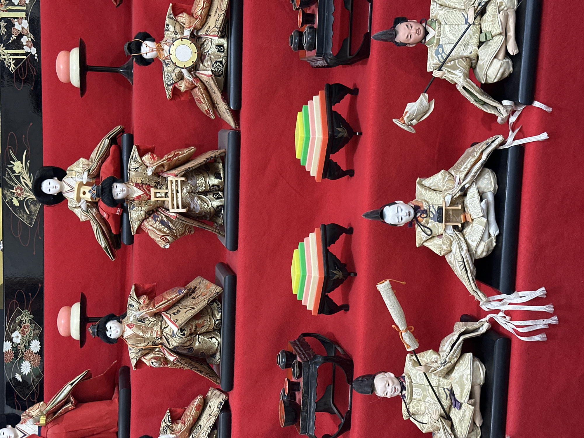The image displays a collection of traditional Japanese Hina dolls, arranged on a red cloth backdrop. The dolls include depictions of the Emperor and Empress, as well as various attendants and accessories like tiered stands and ceremonial objects. The dolls are richly adorned in traditional Japanese garments. The background is a simple red cloth, which provides contrast and emphasizes the dolls. The scene appears to be a display of Hina dolls for the Hina Matsuri festival. This scene could be found in any place where people celebrate Hina Matsuri and is not specific to Quetta City Tehsil, Pakistan.