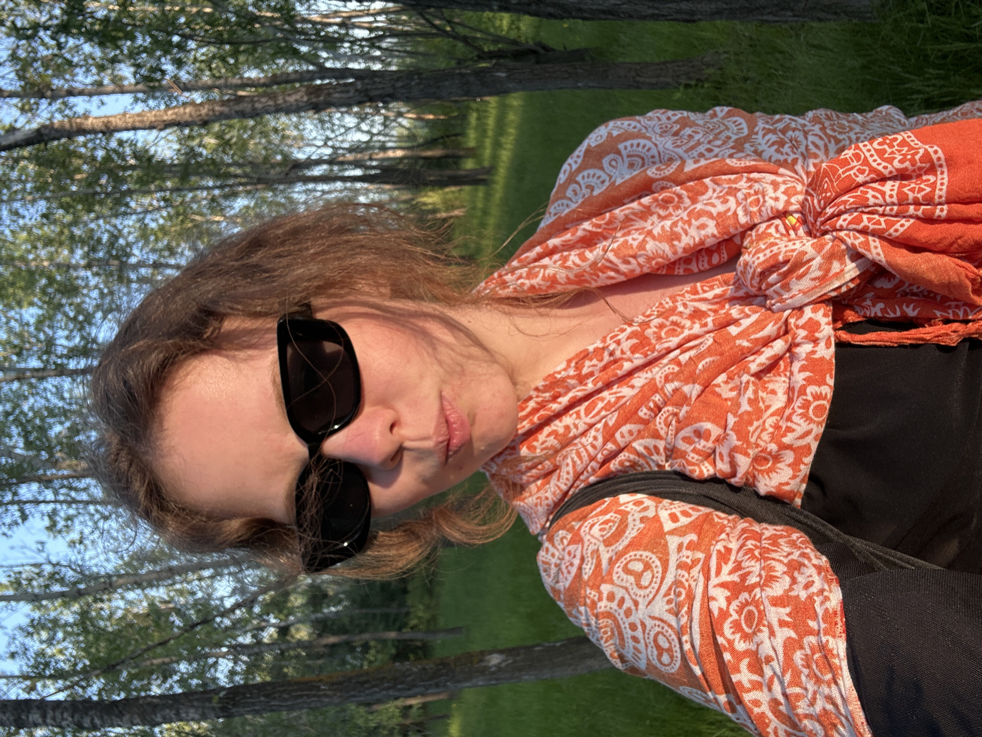A young woman is the central subject of this outdoor portrait, positioned with her head tilted to her left and facing the camera. She has shoulder-length, wavy reddish-brown hair and wears black sunglasses, obscuring her eyes. Her lips are slightly pursed, suggesting a casual or playful expression. She is dressed in a dark top, likely black, and a vibrant orange and white patterned scarf with a paisley or floral design, which is wrapped around her upper body. A dark strap, possibly from a backpack, is visible on her right shoulder.
The background depicts a natural setting with a line of tall trees, featuring slender trunks and green foliage, consistent with a park or wooded area in Krasnoyarsk, Russia. The ground beneath the trees is covered in lush green grass. The scene is illuminated by warm, bright sunlight, indicating late afternoon or early evening, with a clear blue sky visible through the tree branches. No text is visible in the image.