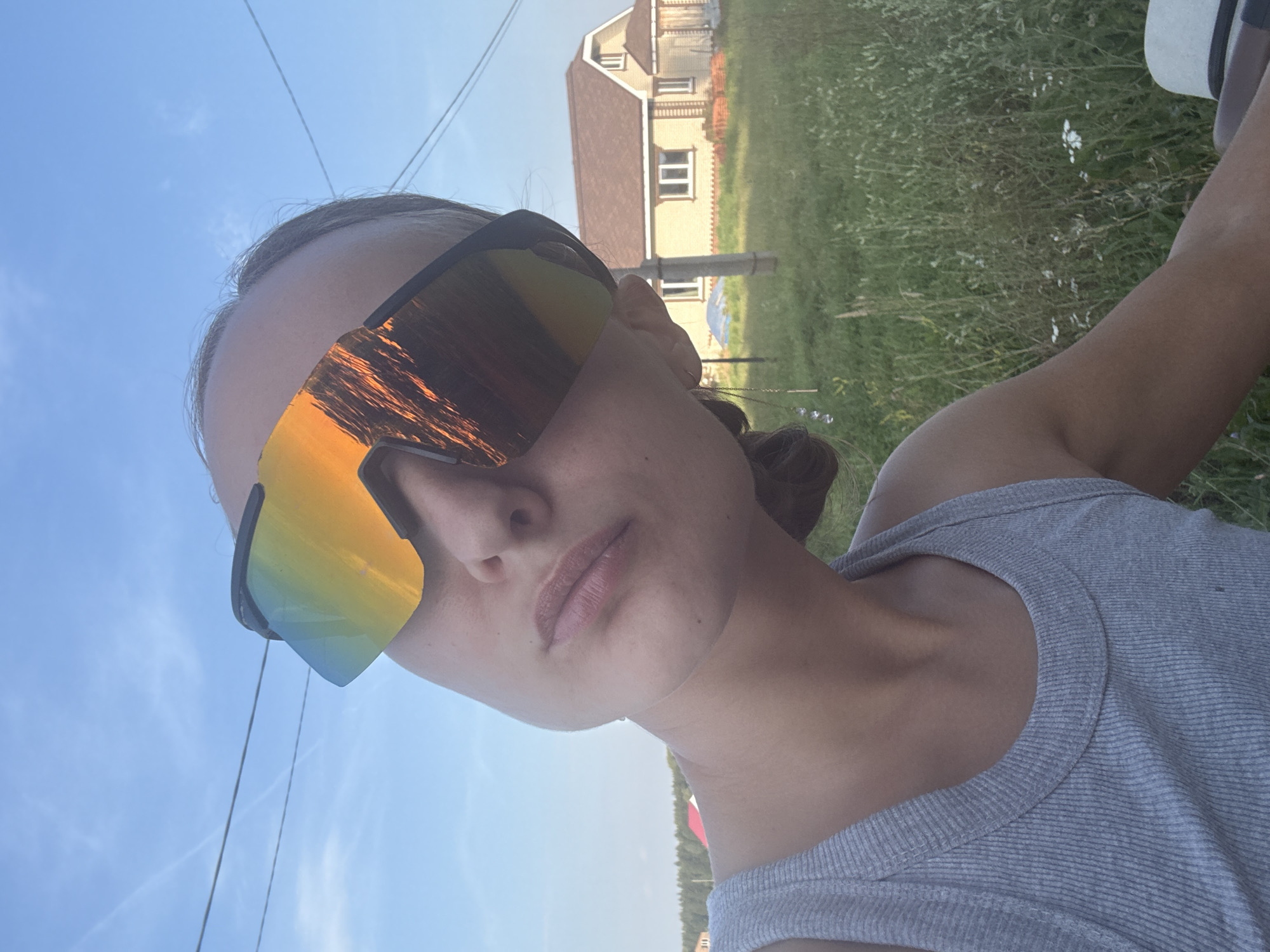 The media file shows a close-up, portrait-style selfie of a young woman wearing large, reflective, multi-colored sunglasses and a grey tank top. The sunglasses reflect a bright sky and possibly some distant trees or a building. The woman's face is angled slightly away from the camera, and her lips are parted. Her hair is dark and appears to be pulled back.
The background is a sunny outdoor scene with a clear blue sky and some wispy clouds. There is green grass and some small white flowers visible. In the mid-ground, a house with a brown roof and yellow siding is visible, as are some power lines. The overall impression is of a warm, sunny day, likely in a rural or semi-rural setting, consistent with the provided location context of Подшивалово, Russia.
The time of day appears to be daytime, judging by the bright sunlight and clear sky. No specific event is depicted, beyond the act of taking a selfie. There is no visible text in the image. The emotion conveyed is casual and perhaps relaxed, as one might feel on a pleasant day outdoors.