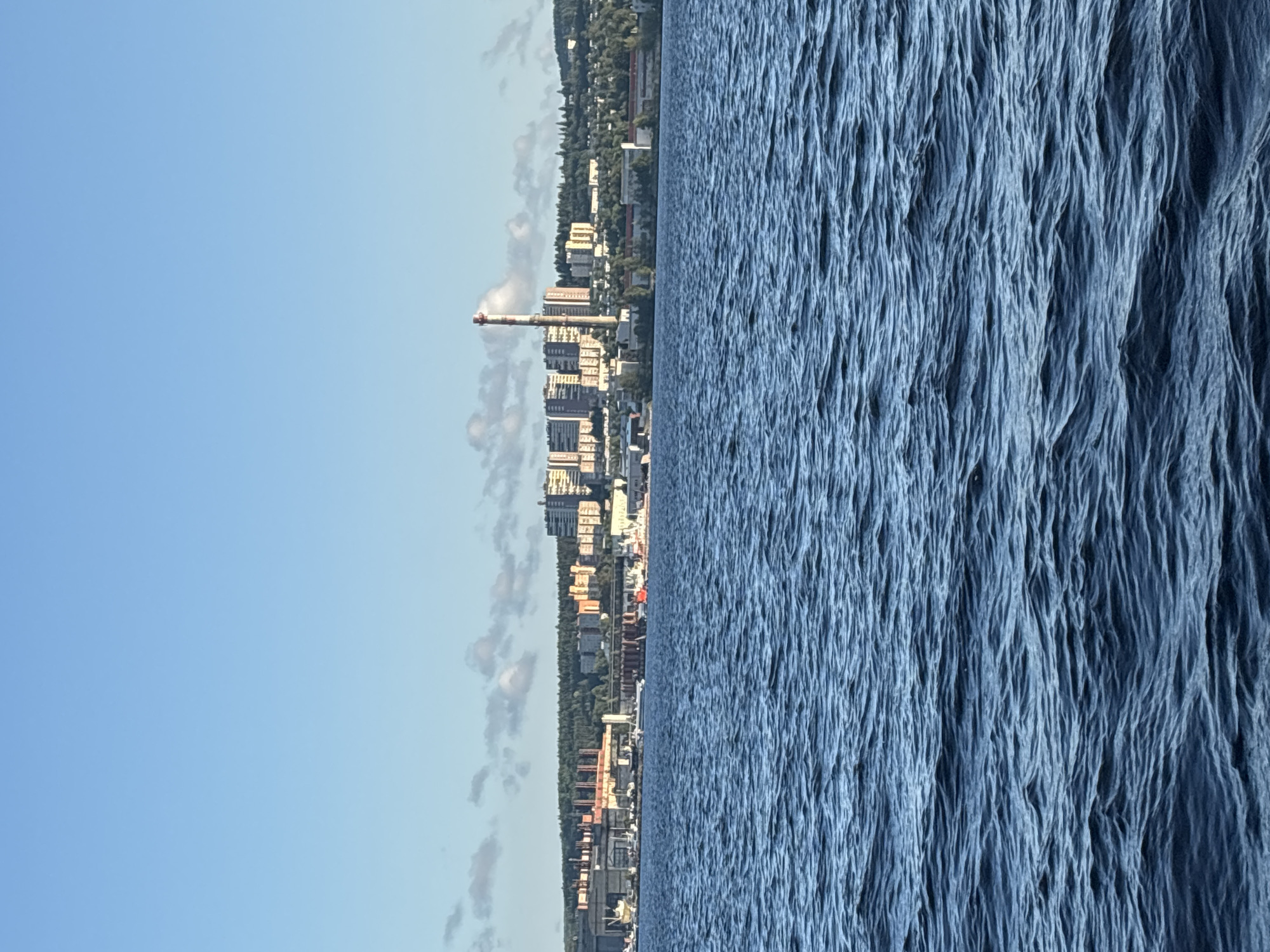 The image shows a panoramic view of the city of Petrozavodsk, Russia, from across a body of water. The foreground is dominated by the deep blue, choppy surface of the water, with small waves creating a textured effect. In the middle ground, the city's shoreline is visible, featuring a mix of industrial buildings, apartment complexes, and scattered trees. A tall, red and white striped smokestack stands out prominently among the buildings. Several boats and ships are docked along the waterfront. The background consists of a clear blue sky with a few scattered white clouds. The overall scene depicts a coastal industrial city under a bright sky.