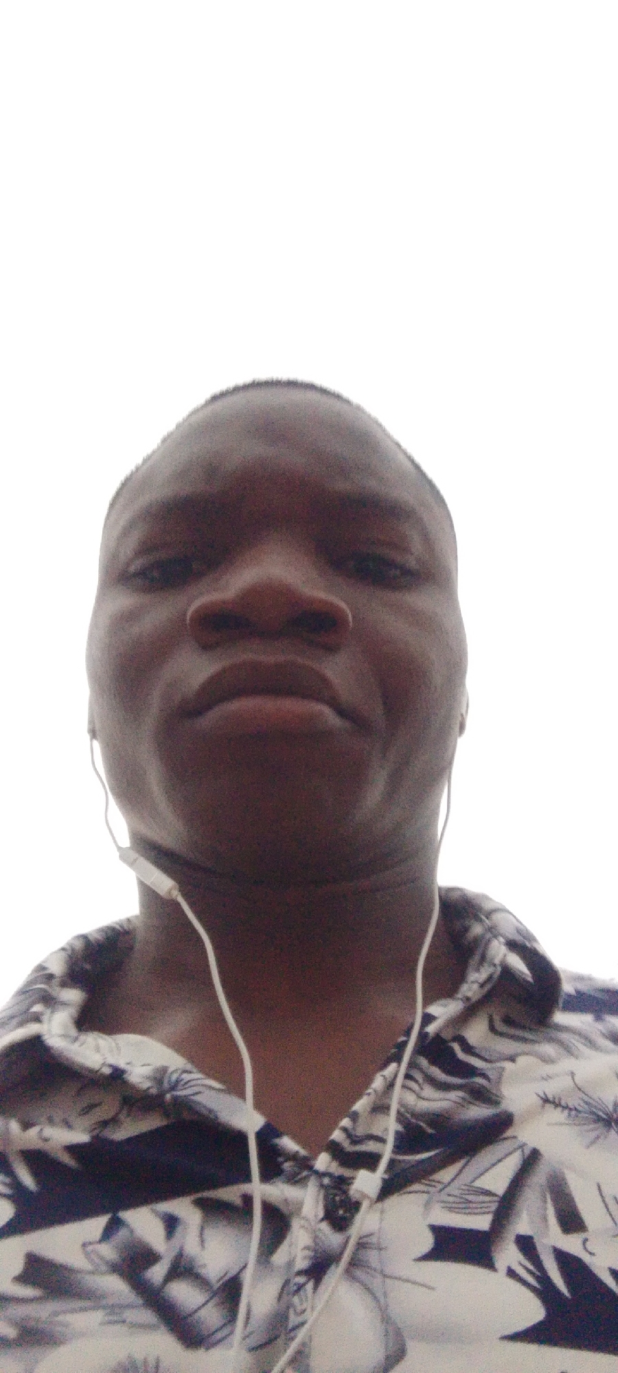 A close-up, low-angle shot shows a person's face, wearing earphones and a patterned shirt. The subject is looking directly at the camera with a neutral expression. The background is plain white, indicating it may be an indoor setting or an outdoor setting with an overcast sky. The subject's skin tone is dark, and their hair is short. The shirt has a black and white floral or abstract pattern. The earphones are white and appear to be wired. The lighting is even, with no harsh shadows.