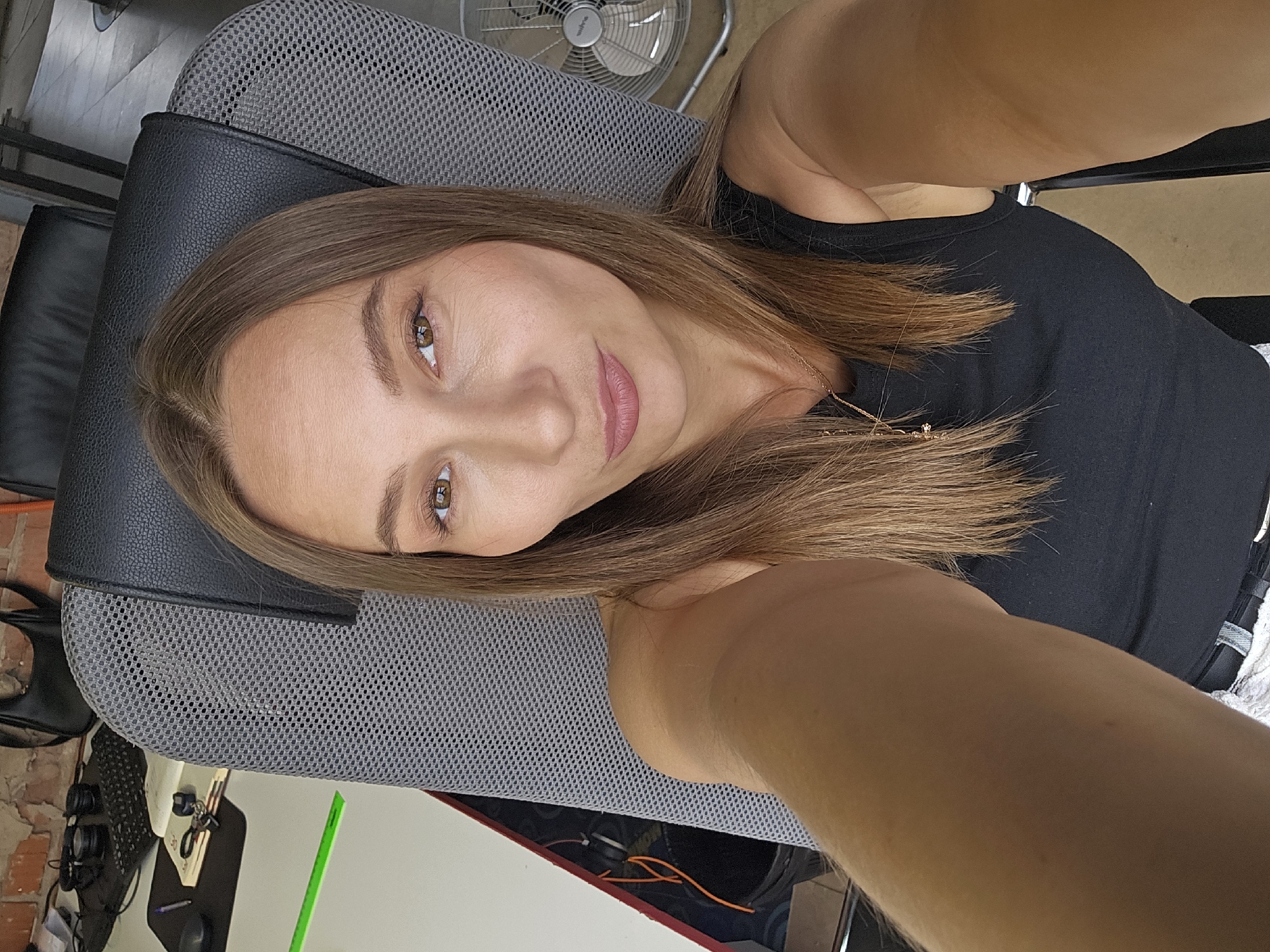 A person is taking a selfie. The person has medium-length brown hair, light skin, and is wearing a black tank top. The person is sitting in a gray mesh office chair. In the background, there is a white desk with a green stripe, a black leather chair, a floor fan, and part of a brick wall. The location in the city of Chaikovsky, Russia cannot be determined from the image.