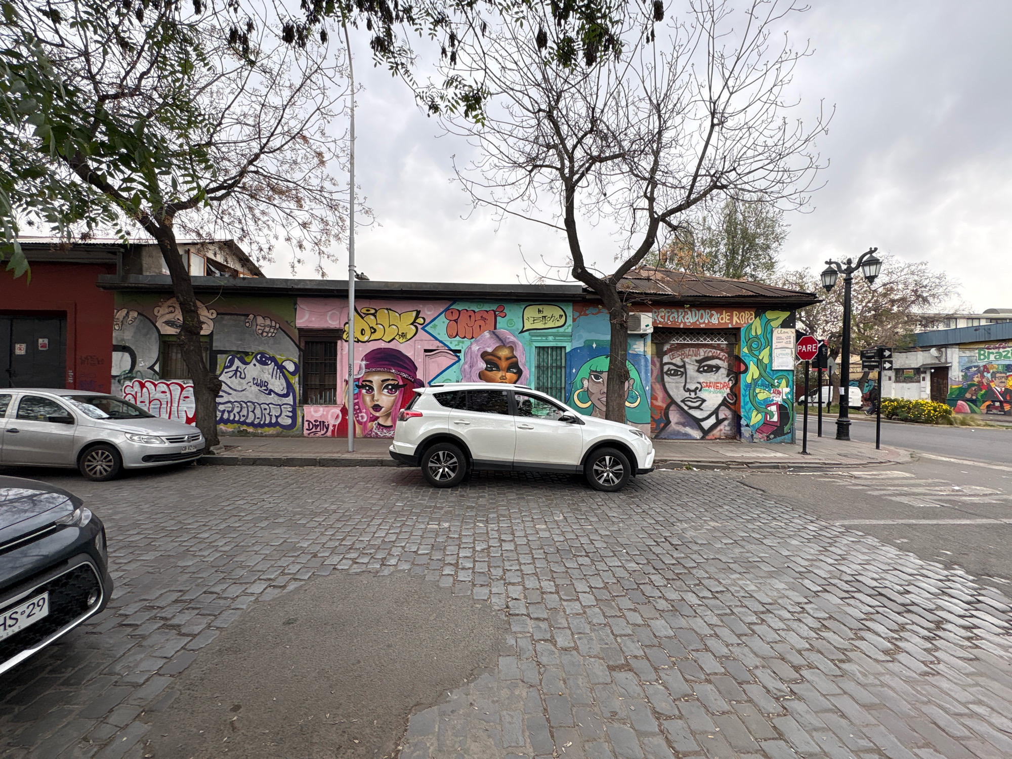 The image captures a street scene in Santiago, Chile, dominated by vibrant graffiti art on the facades of buildings and a cobblestone street. A white SUV is parked on the side of the street, aligned with a row of buildings adorned with colorful murals of faces and abstract designs. A silver sedan is parked further down the street. The architecture appears to be a mix of older, single-story structures with corrugated metal roofs and some newer additions. Bare trees with intricate branches are visible, suggesting a cooler season. A street lamp stands on the right side of the frame, and a "PARE" (Stop) sign is mounted on a pole near an intersection. The sky is overcast, contributing to a diffused, natural light. The cobblestone street shows varying textures and some wet patches, indicating recent rain or dampness. The overall impression is one of an urban environment that embraces street art as a significant part of its visual identity.