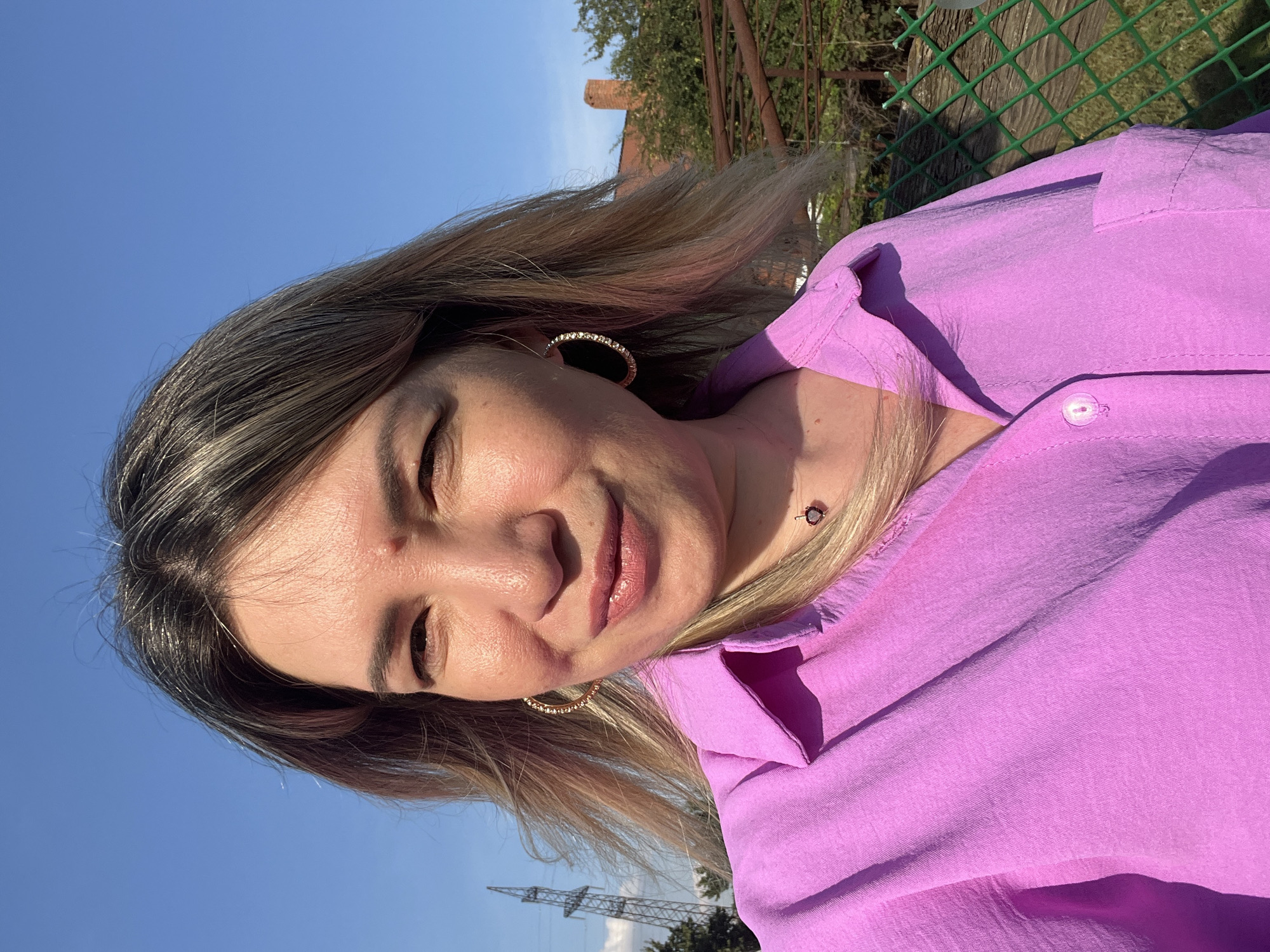 A woman with blonde hair is taking a selfie outdoors. She is wearing a pink collared shirt and large hoop earrings. The background shows a blue sky, trees, a fence, and a tall industrial tower with power lines. The woman is looking at the camera and smiling slightly. The location is identified as Yanovo-Grushevsky, Russia.