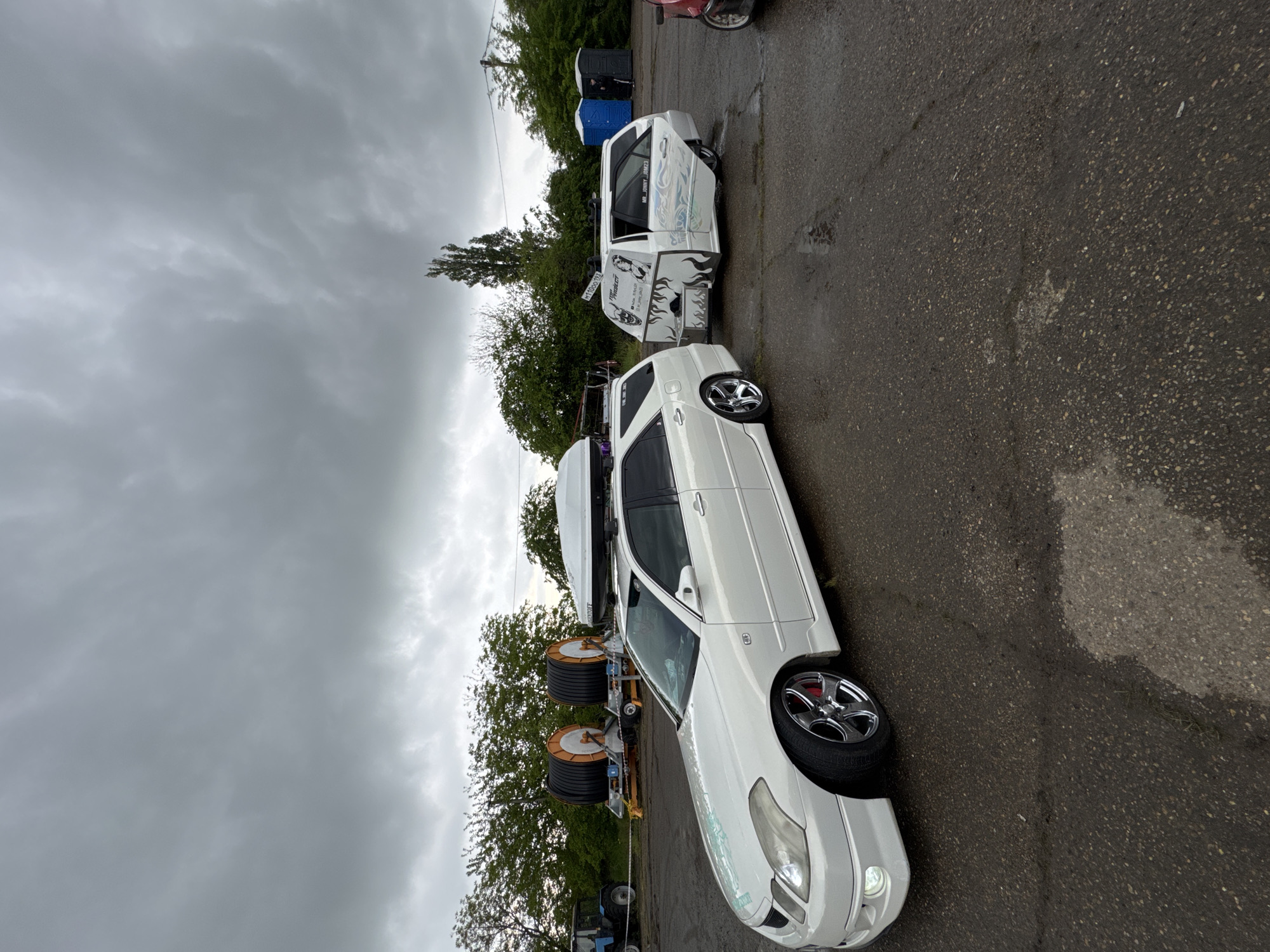 The image depicts two white vehicles parked on an asphalt surface, likely a parking lot. The sky is overcast. The vehicles are a white station wagon with a cargo carrier on top and a white truck with customized art. Utility cables on a trailer can be seen in the background along with portable toilets. The location is the city of Pavlovskaya, Russia.