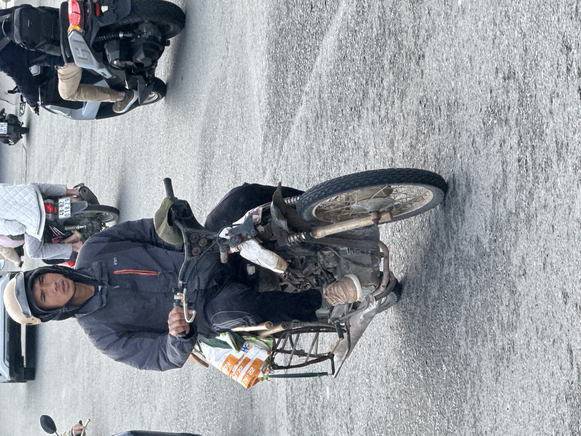 A man wearing a tan helmet and a dark jacket rides a motorcycle on a paved road in Da Lat, Vietnam. The motorcycle has a metal rack on the back carrying two white and orange bags. The man is looking forward, holding the handlebars. Other vehicles, including another motorcycle and a car, are visible in the background. The road surface is asphalt with some textured areas.