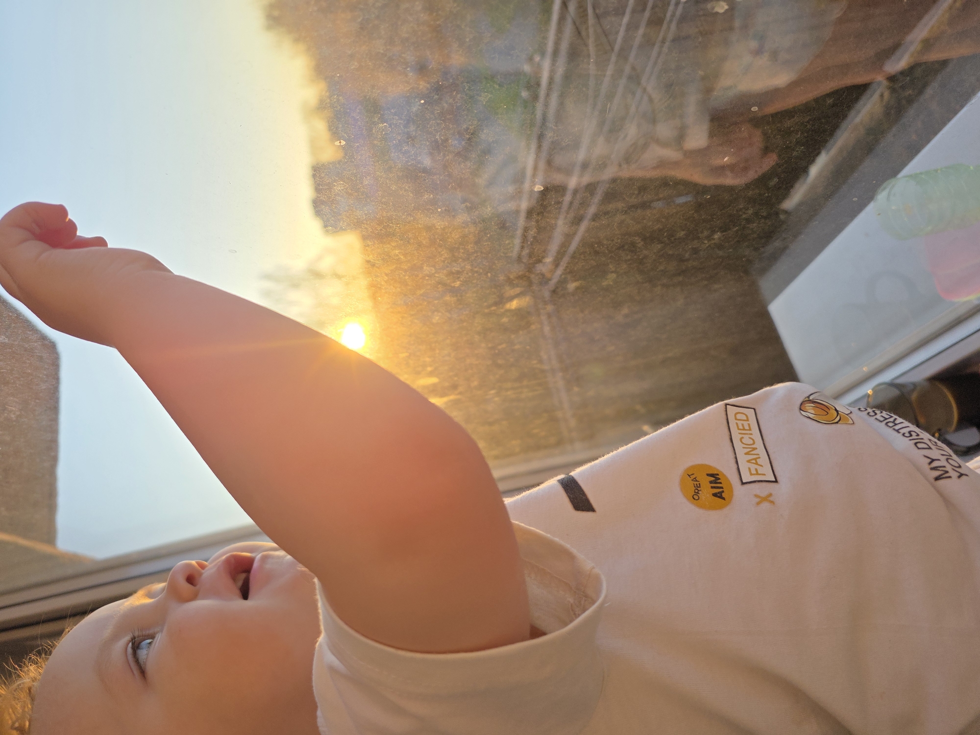 A low-angle, rotated view depicts a child lying down, possibly near a window. The child's arm is raised, reaching towards the light filtering through the dirty glass. The reflected sunlight creates a bright, diffused effect. The child's shirt has printed text and a design. The location is potentially Chaikovsky, Russia based on the user's prompt. Other objects are indistinct behind the dirty window.