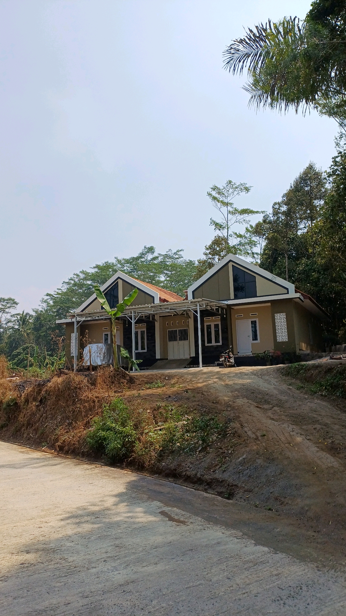 The image shows a scene in Kebumen Krajan, Indonesia, featuring two adjacent, modern-style houses situated on a slope. The houses have light brown and dark gray facades, with distinctive angular roofs topped with reddish-brown tiles. Each house has a covered porch supported by white metal frames. The house on the left has white-framed windows and a covered area with a white tarp. A banana tree is planted in front of the left house, and a motorcycle is parked in front of the right house. The houses are surrounded by lush green trees and vegetation, with a dirt and concrete road in the foreground. The sky is clear and pale. The terrain is uneven, with a visible slope leading up to the houses.