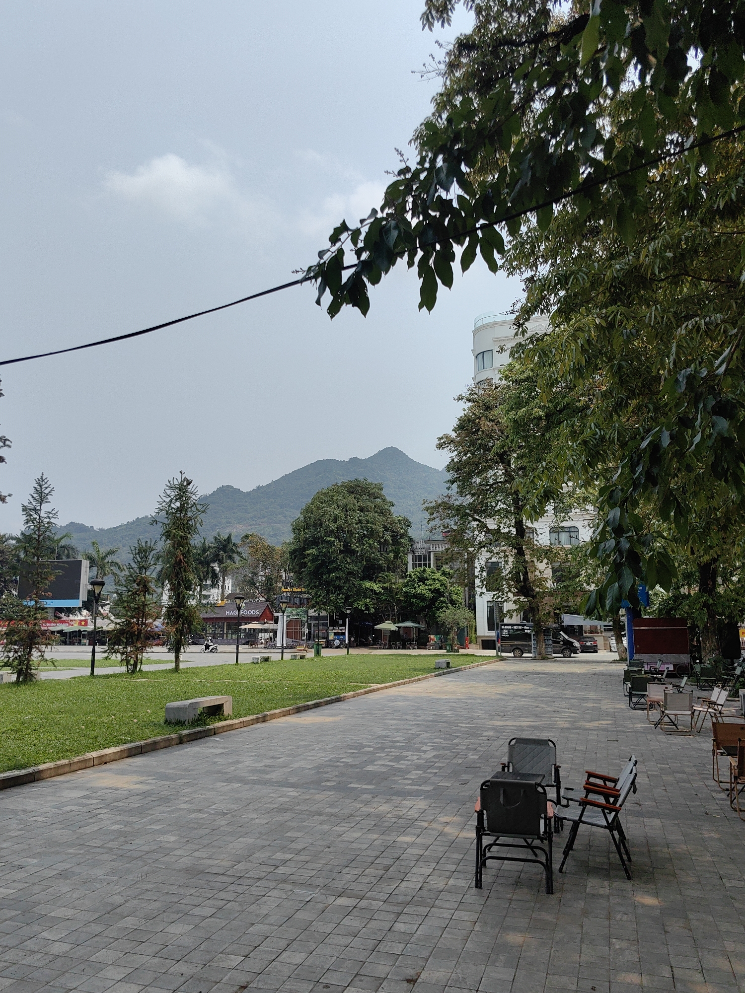 The photograph shows a paved area in Thành phố Hà Giang, Vietnam. Two sets of folding chairs are arranged on the paved surface. A strip of grass separates the paved area from a street with various commercial establishments, including a restaurant named "HAG FOODS". Beyond the street, a mountain range forms the backdrop. Trees are present throughout the scene, partially obscuring buildings and adding to the natural environment. The sky is overcast.