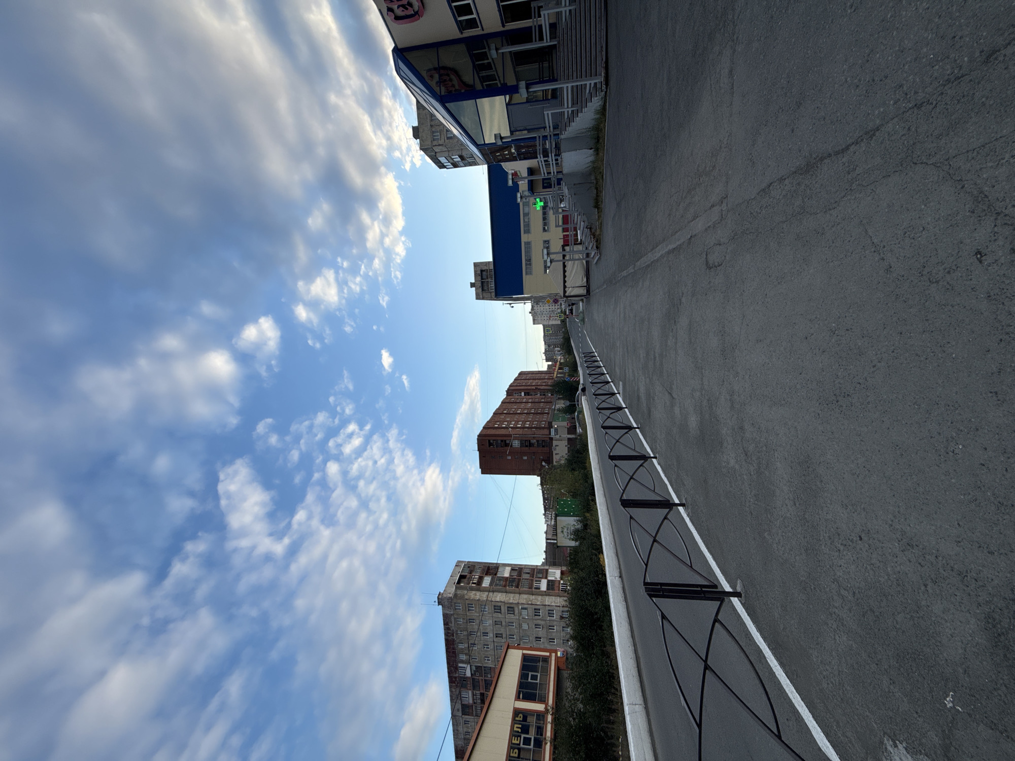 The image depicts a wide asphalt street in Norilsk, Russia, bordered by a black metal fence with an ornate, wavy design on the left side. The street appears to be a sidewalk or pedestrian walkway. On the right side of the street, there are buildings. The closest building on the right is a modern structure with a blue roof and large glass windows, featuring a prominent green cross sign, likely indicating a pharmacy. Further down the street, there are other buildings, including a long, dark red brick building and several grey, utilitarian apartment blocks with visible balconies. The sky above is a mix of blue with scattered white and grey clouds, suggesting it may be daytime with variable weather. The overall impression is of a typical urban street scene in a northern Russian city.