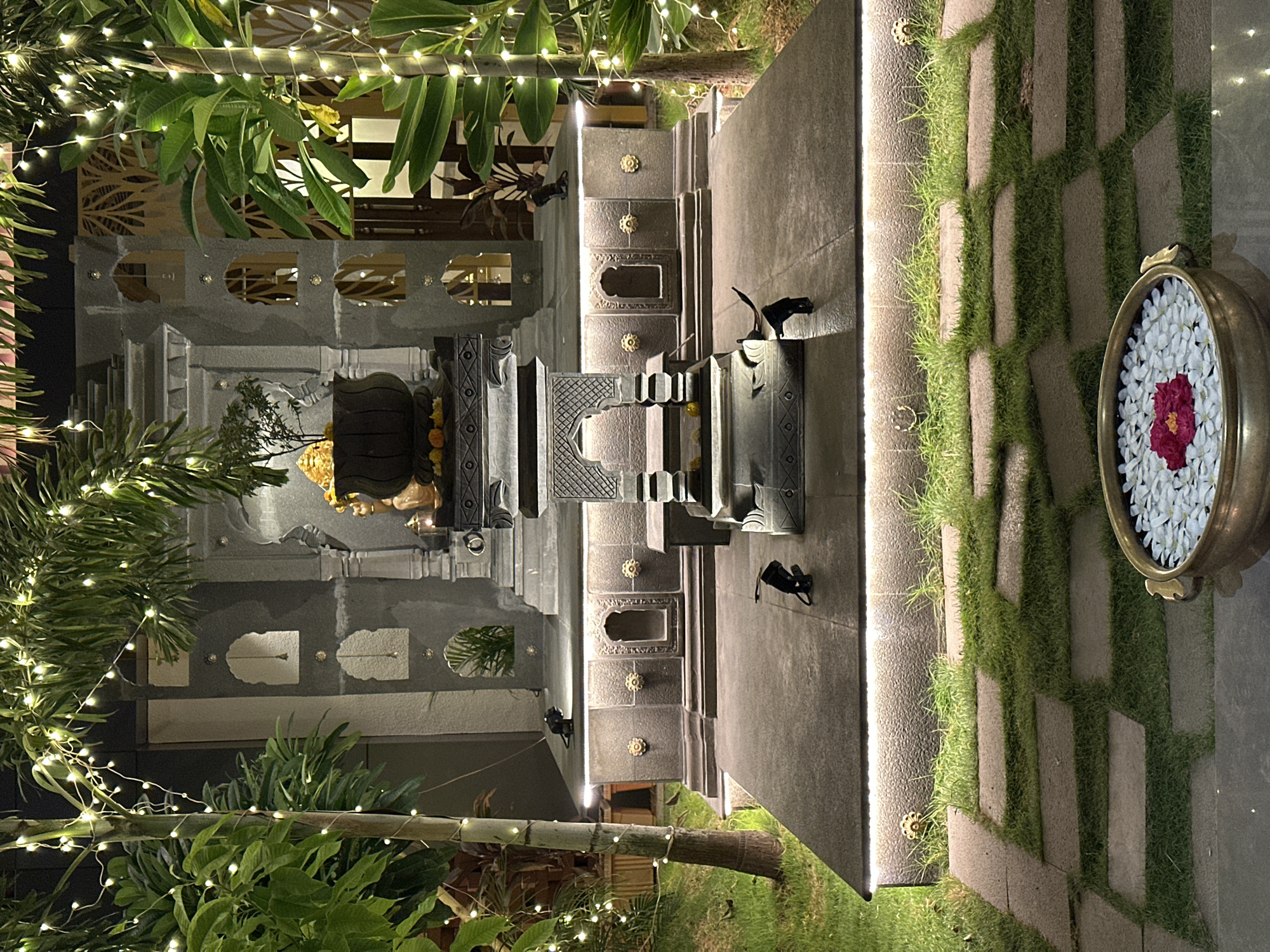 The image shows a courtyard or garden area at night, likely in Kalamb, India, given the architecture and cultural elements. The scene is well-lit with string lights and spotlights. The focal point is an ornate, multi-tiered stone structure resembling a Hindu temple or altar. It has detailed carvings and is decorated with flower garlands.
To the right of the structure is a pathway paved with stone slabs and patches of grass. A brass bowl filled with white and red flowers sits near the edge of the pathway. Lush greenery surrounds the area, and the scene has been decorated with lights. The image seems to capture a tranquil, celebratory atmosphere.