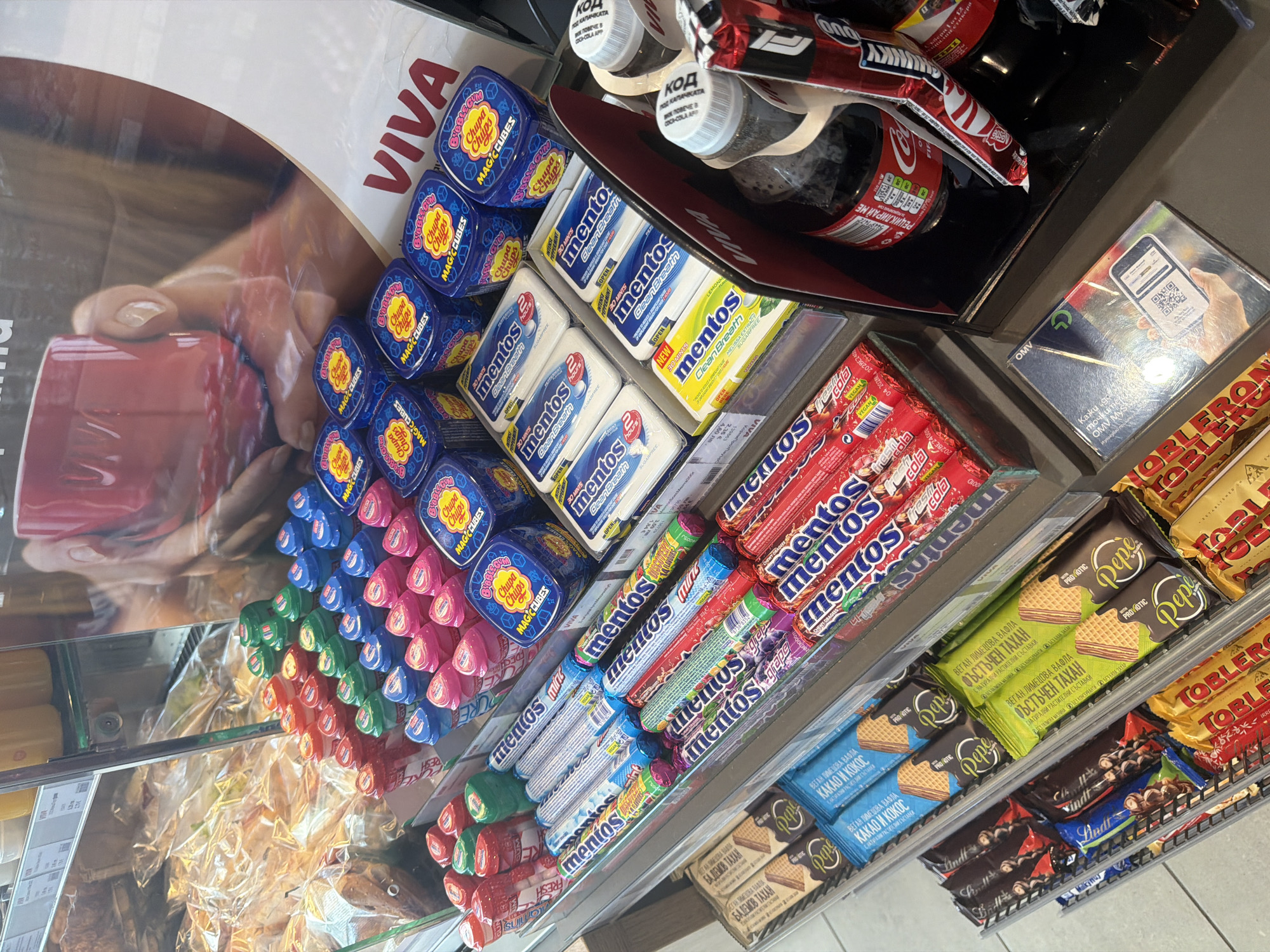 This image captures a well-stocked retail display within what appears to be a convenience store, likely at an OMV gas station, given the "OMV MYS" branding on a sign. The location is Borovo, Bulgaria.
The shelves are filled with various confectionery items. Prominently featured are rows of Chupa Chups Magic Cubes bubble gum in blue, pink, green, and red packaging. Below these, there are several flavors of Mentos Clean Breath mints in white and yellow packaging, as well as rolls of Mentos in different flavors like cola, grape, fruit mix, and mint.
Further down, a section displays "Pepe" brand vegan wafers, with flavors such as "Cacao and Coconut", "Almond Tahini", and "Peanut Tahini", indicating natural plant-based ingredients. Adjacent to these are classic Toblerone chocolate bars and a selection of Lindt chocolates.
In the upper right, Coca-Cola bottles are visible, some with white caps bearing Cyrillic text "КОД" (code) and "ВОК ПОВЕЧЕ В КОКА-КОЛА АРР" (Scan more in Coca-Cola App), suggesting a promotional campaign. One bottle also has "РЕЦИКЛИРАЙ МЕ" (Recycle me) printed on it. A prominent display banner for "VIVA" features a close-up of hands holding a red mug.
A small, reflective sign shows a QR code with text "OMV MYS", "Kaku go", and "точките" (points), along with a numerical code, hinting at a customer loyalty program or digital payment option. Price labels indicate items are sold in Bulgarian Lev (лв.) and Euro (€), with one item priced at "4,60 лв." and "2,35 €". The overall scene suggests a typical snack and beverage section designed for quick purchases.