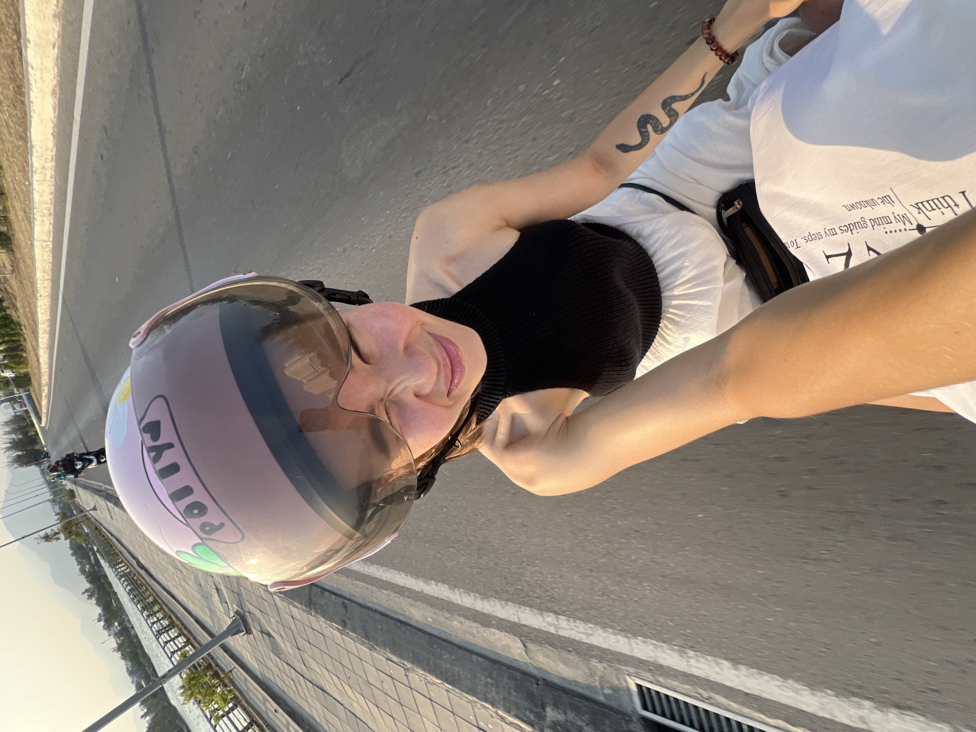 A person is riding a motorcycle, captured at eye-level, in an urban environment. The rider wears a black top, white pants, and a pink helmet with the name "Polly" on it. The rider has a snake tattoo on their arm and a bracelet on their wrist. Another person, presumably the driver, is partially visible, wearing a white t-shirt with a visible phrase "I think." The photograph appears to have been taken in motion. The background reveals a tree-lined roadside bordering a body of water with street lamps and railings. The visible city landscape suggests the location could be near the Nam Natural Heritage Area Trang, Vietnam. The image is a blend of close-up and landscape, suggesting a snapshot taken during a motorcycle ride.