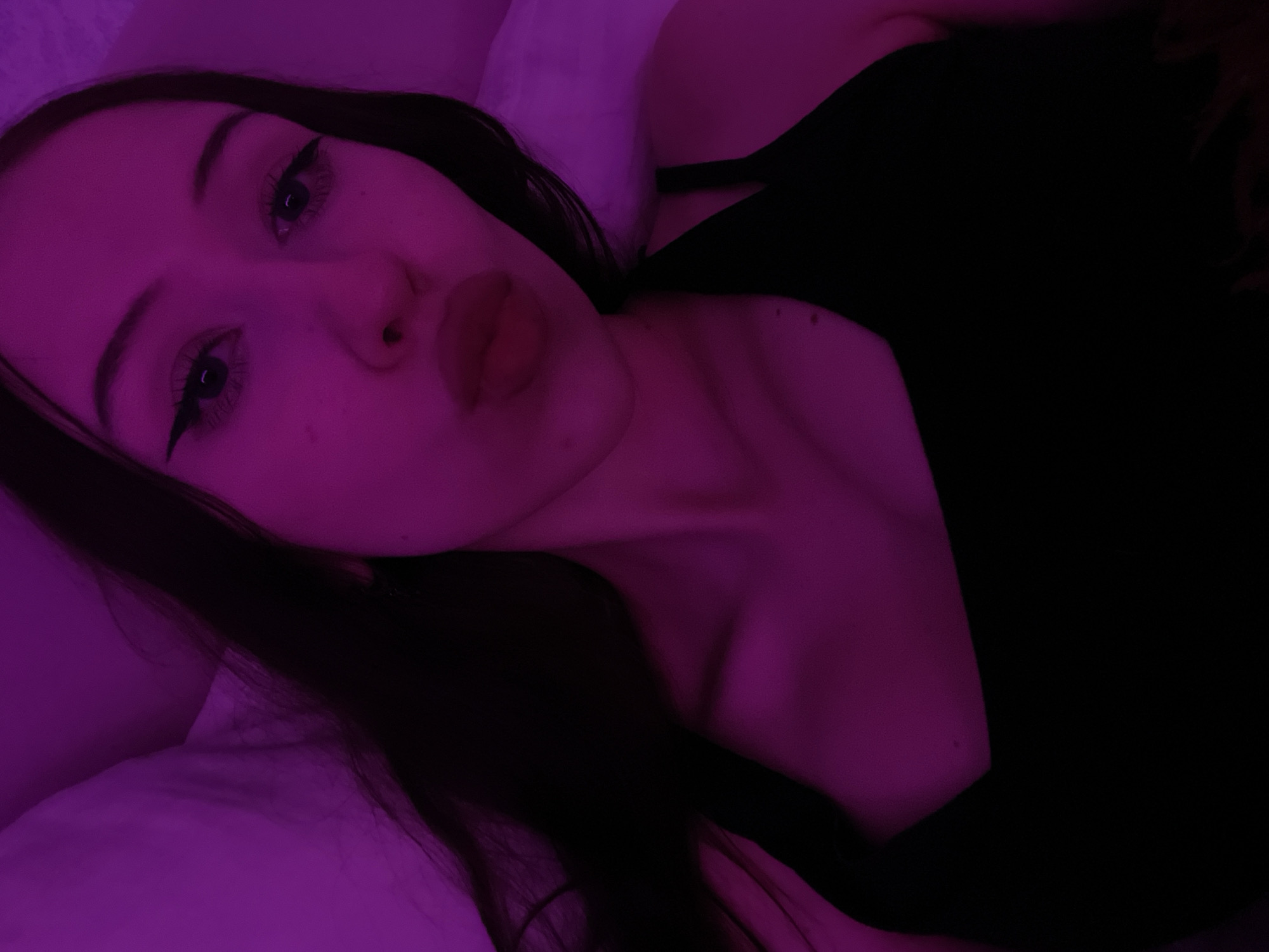 The image is a close-up selfie of a young woman with long, dark hair, wearing a black tank top. She has winged eyeliner and her lips are pursed. The scene is lit with purple light, creating a moody atmosphere. The background appears to be a bed with purple bedding. There is no information in the image to suggest a location in Votkinsk, Russia.